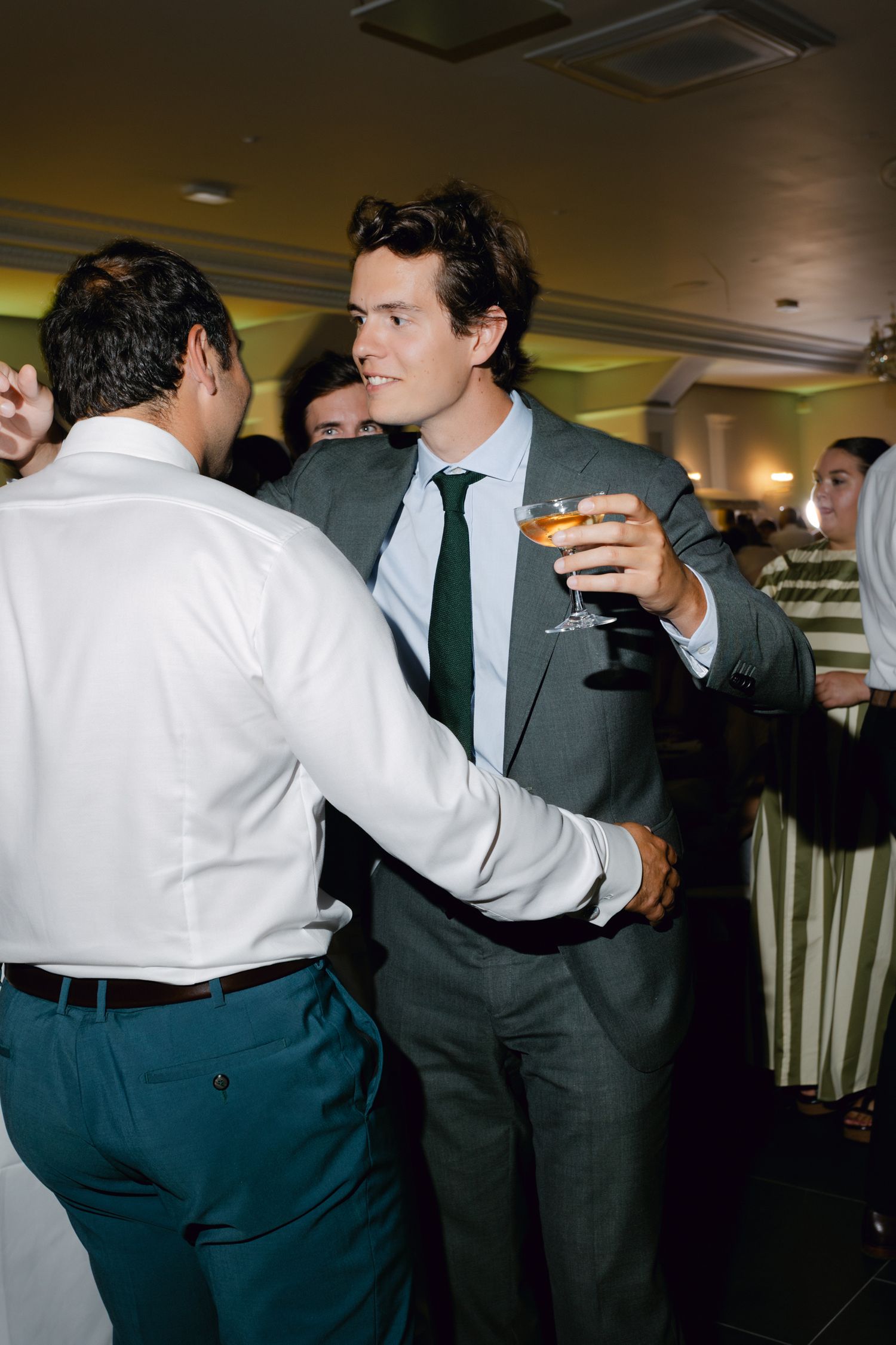 Invité de mariage élégant en costume gris et cravate verte tenant un verre à cocktail tout en socialisant à la réception.