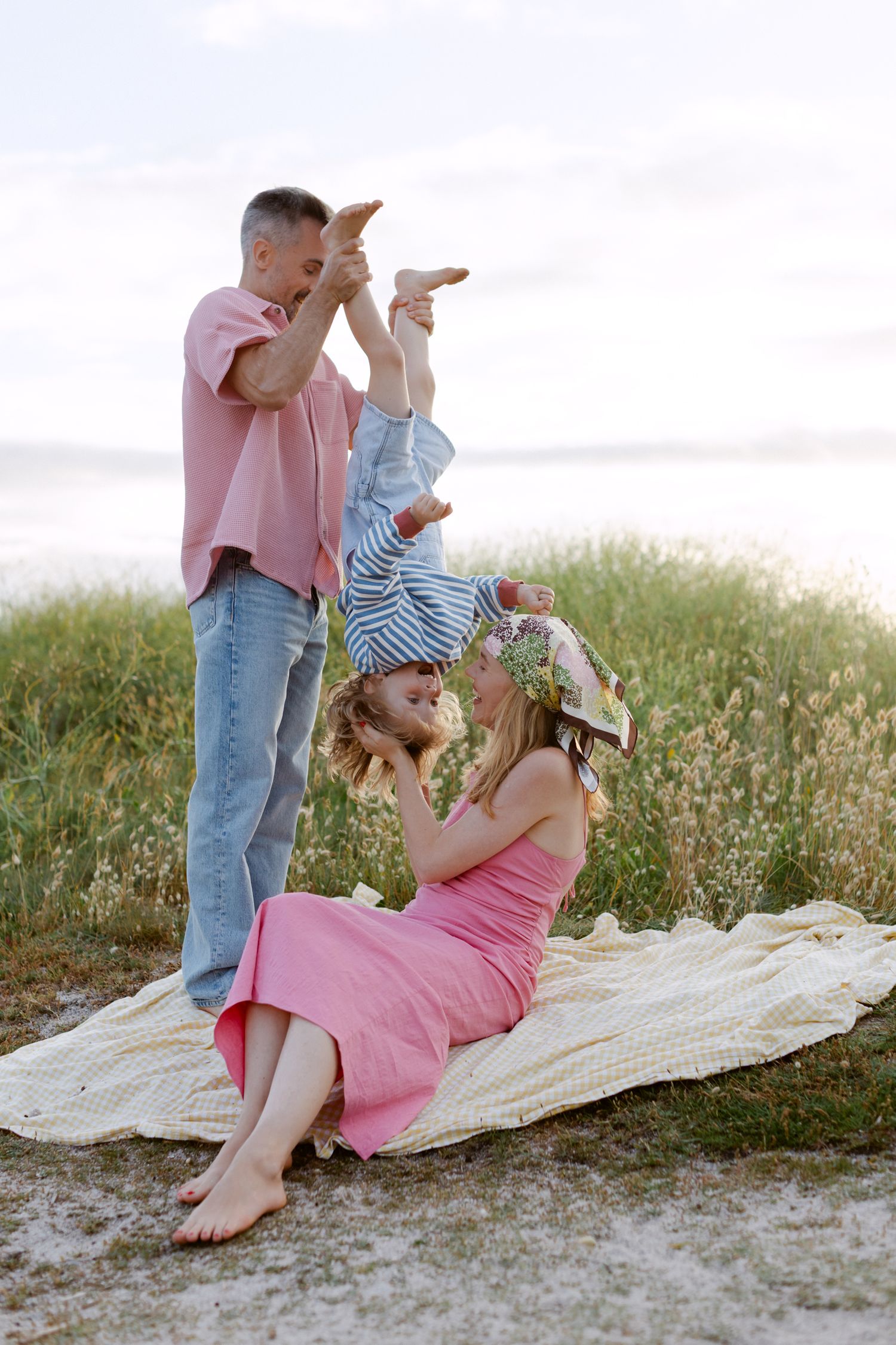 Une famille profite d'un pique-nique sur la plage, papa soulevant un enfant à l'envers tandis que maman est assise sur une couverture.