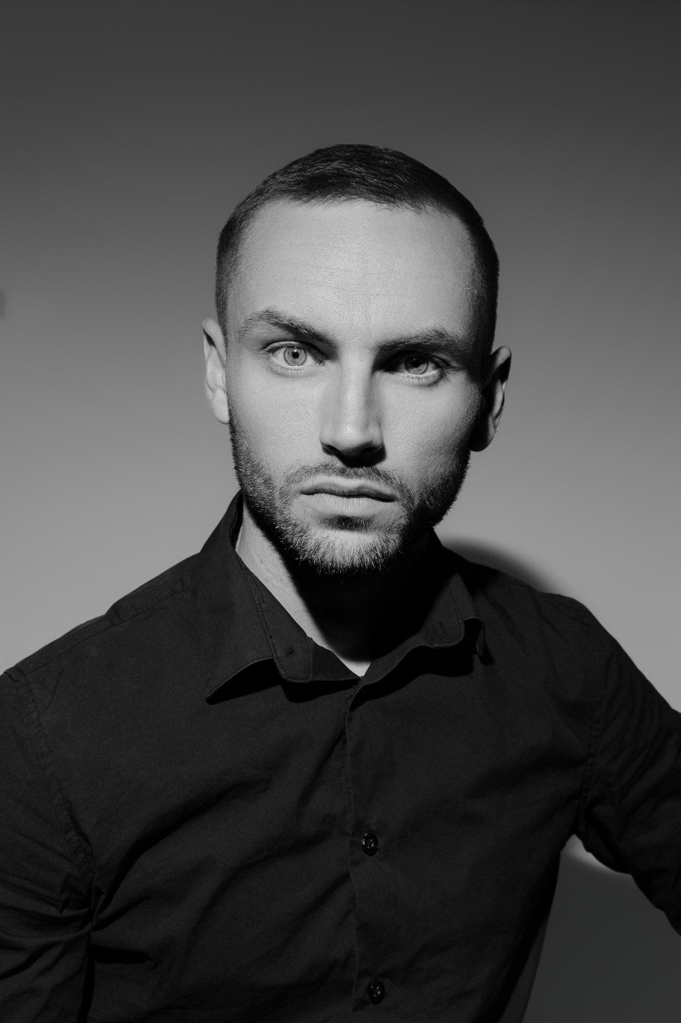 Portrait en noir et blanc d'un jeune homme aux cheveux courts et à la barbe portant une chemise sombre, regardant directement la caméra.
