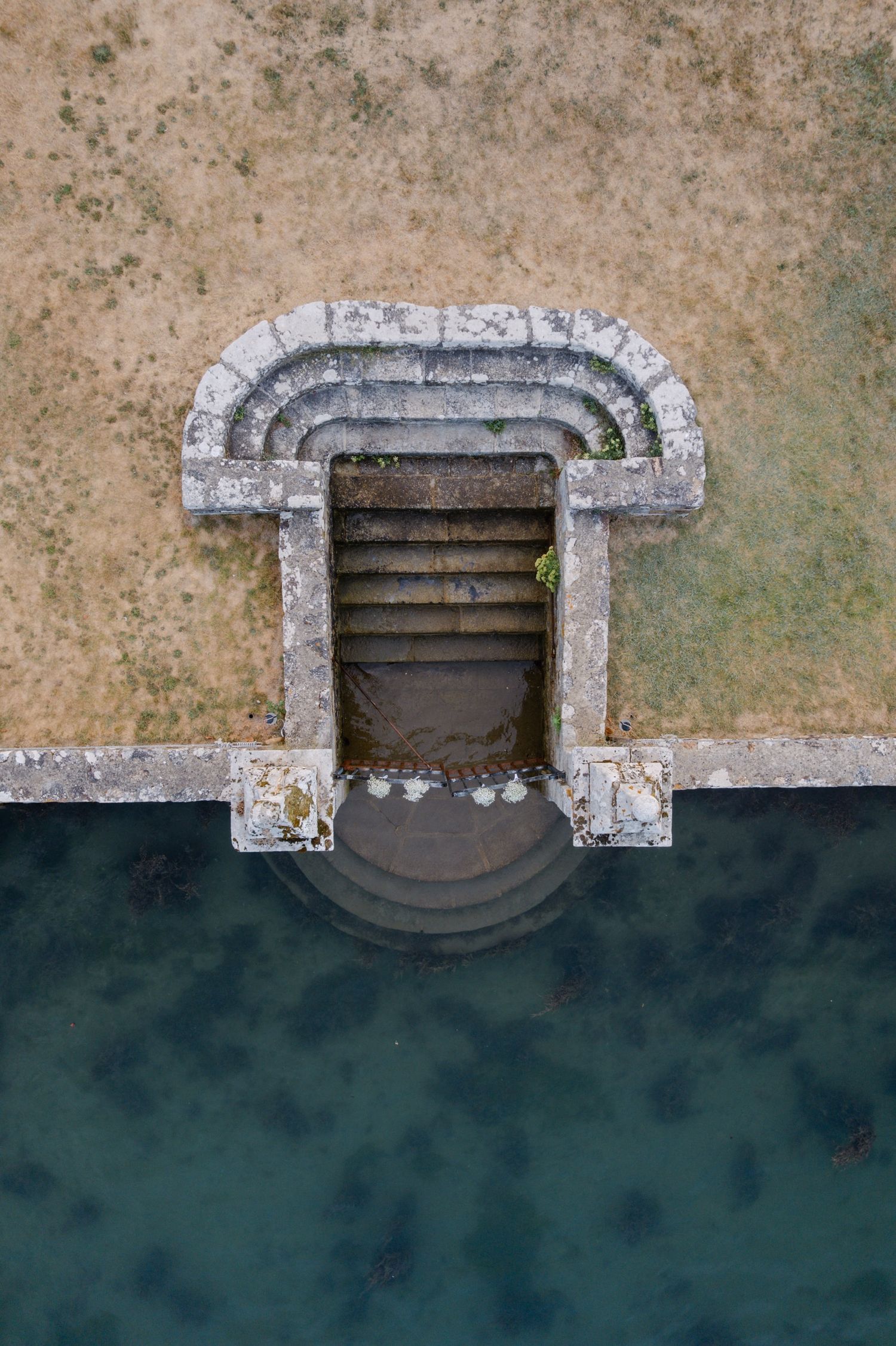Aerial view of ancient stone steps leading into calm green water, framed by a weathered arched stone entrance on grassy ground.