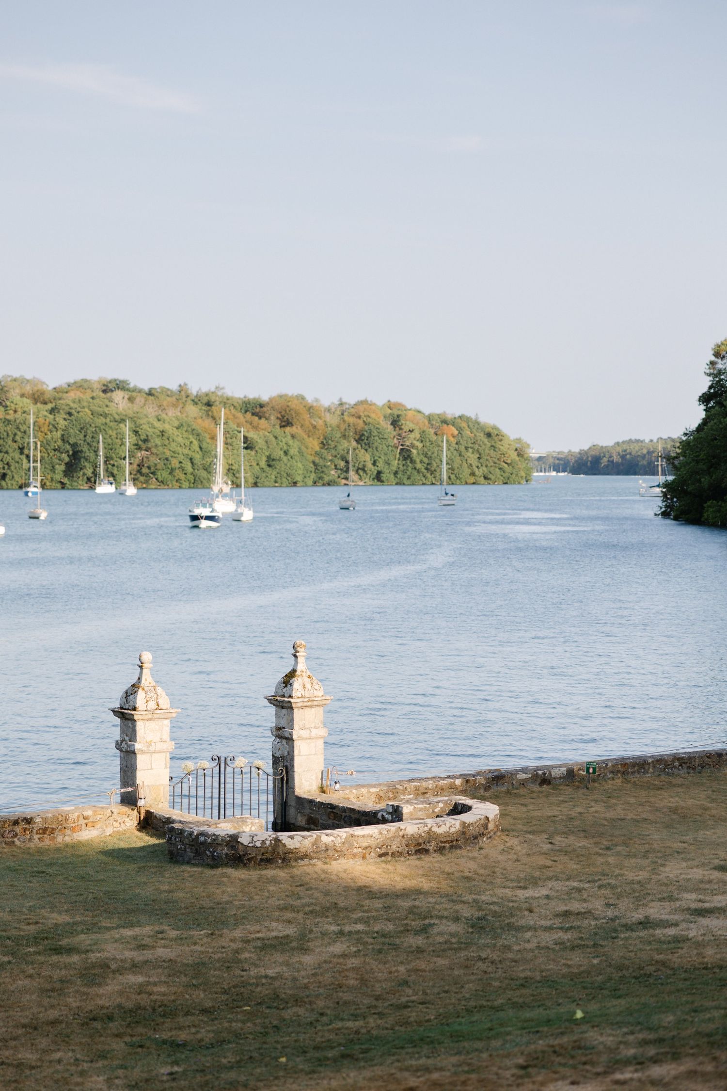 Stone gate pillars beside a calm river with sailboats and lush tree-lined shores under a clear blue sky.