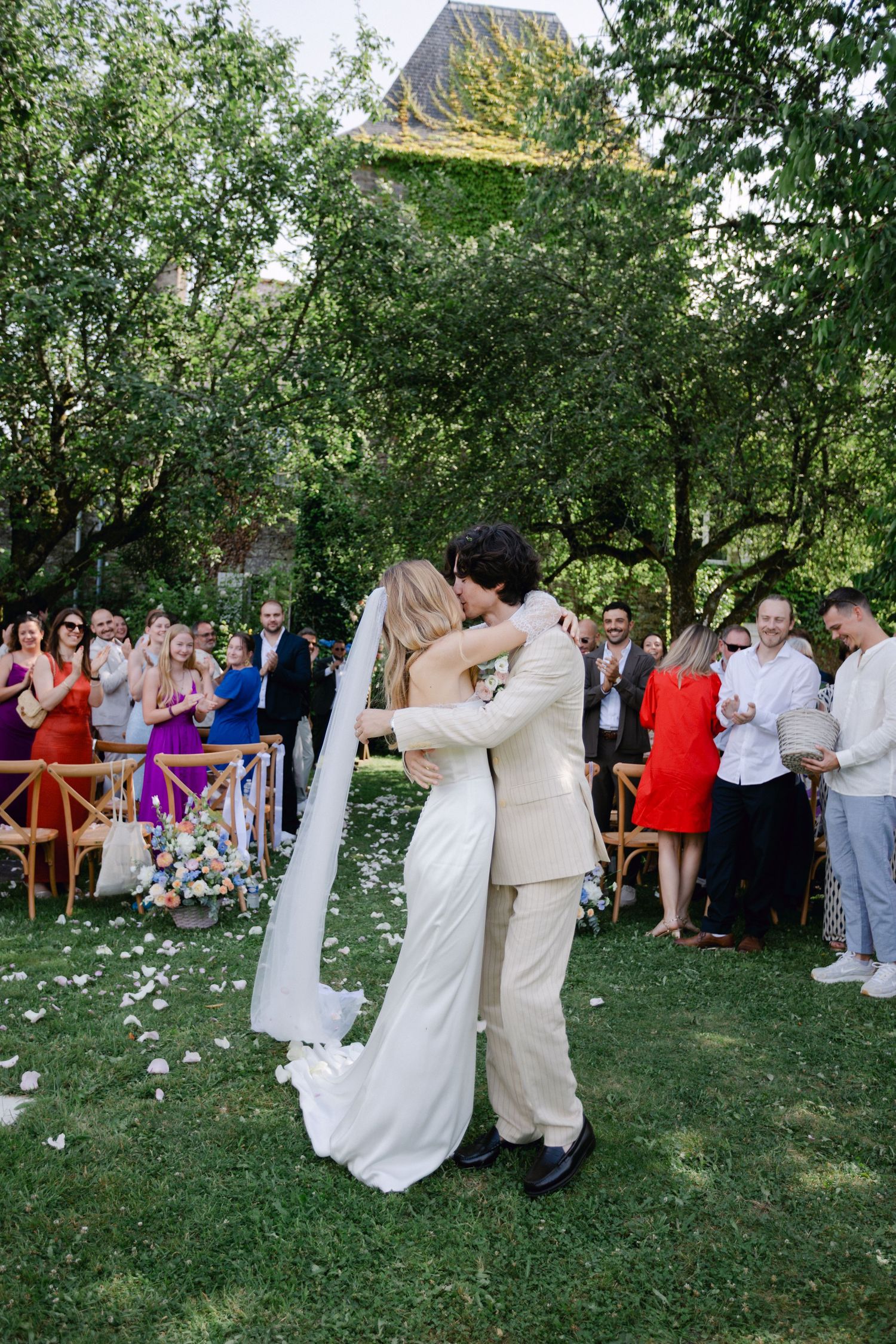 La mariée et le marié partagent le premier baiser sur une pelouse verte entourée d'invités au mariage applaudissant à l'extérieur.