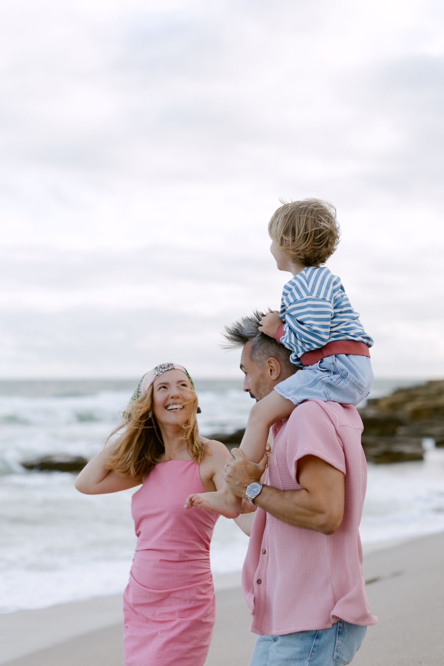 Une famille heureuse à la plage, avec un homme en rose portant un bambin sur ses épaules à côté d'une femme souriante en robe rose.