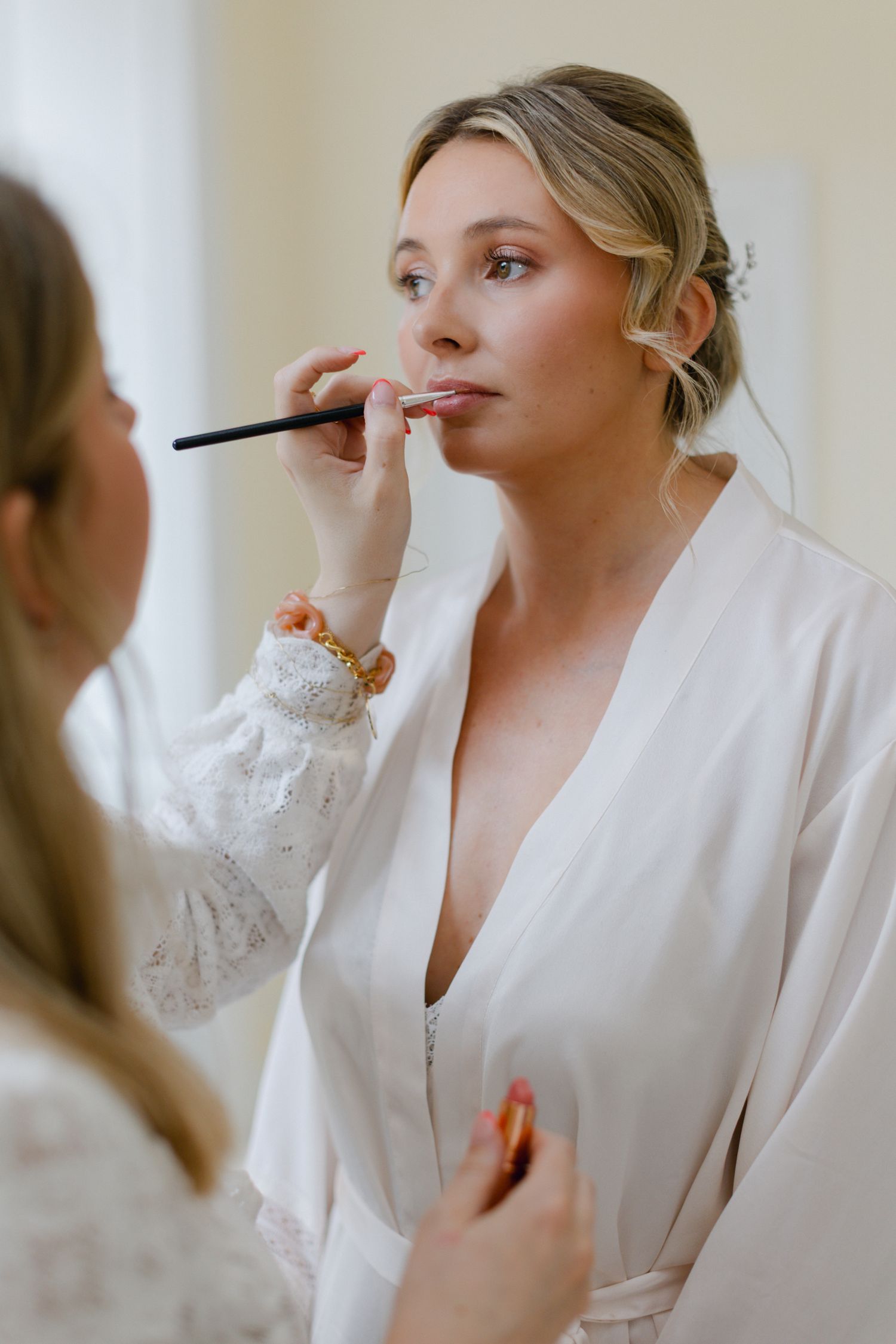 Maquilleuse appliquant du rouge à lèvres à une mariée blonde portant une robe blanche pendant les préparatifs du jour du mariage.