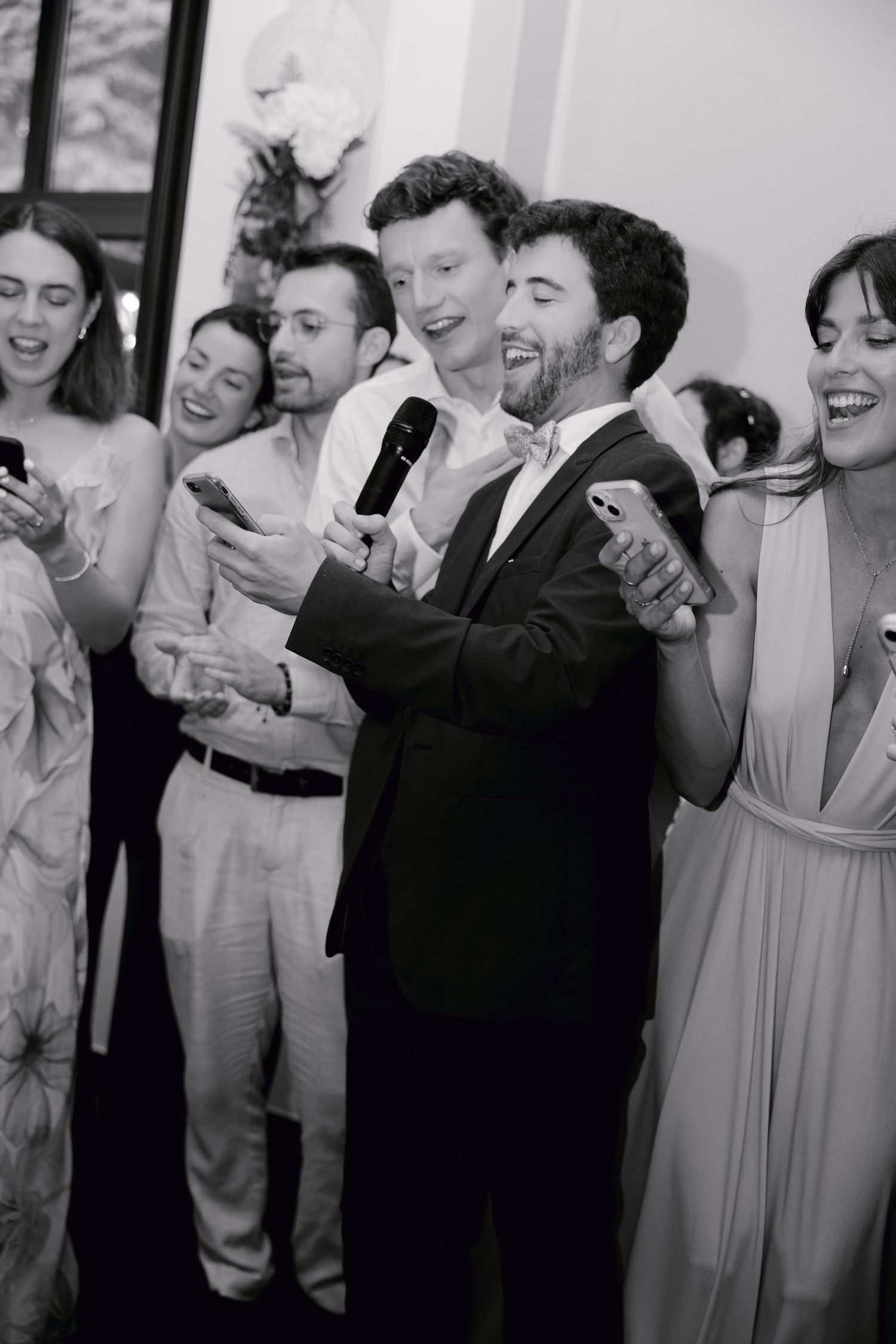 Photo en noir et blanc d'invités riant et chantant avec un microphone lors d'une réception de mariage animée.