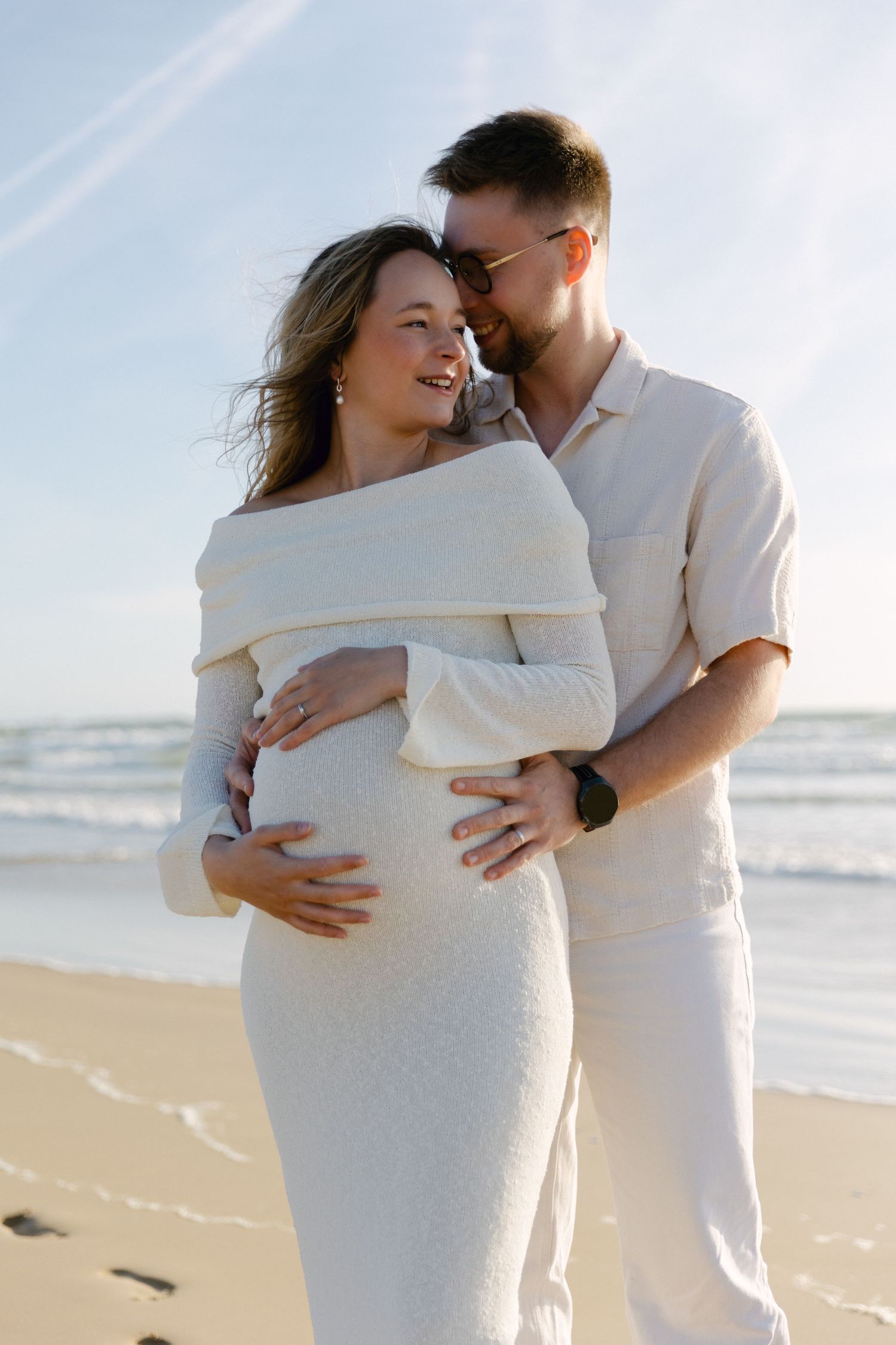 Couple en tenue blanche s'embrassant sur une plage ensoleillée, un homme tenant le ventre d'une femme enceinte près des vagues de l'océan.