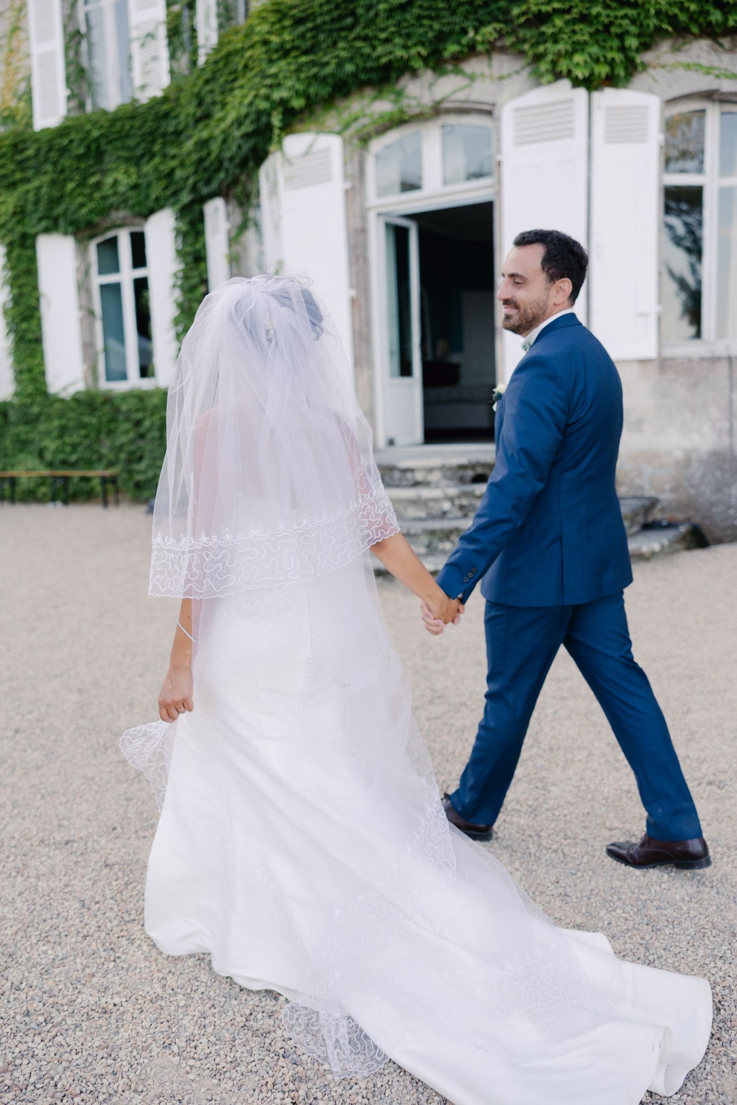 Bride in white gown and veil holds hands with groom in blue suit walking toward ivy-covered venue.