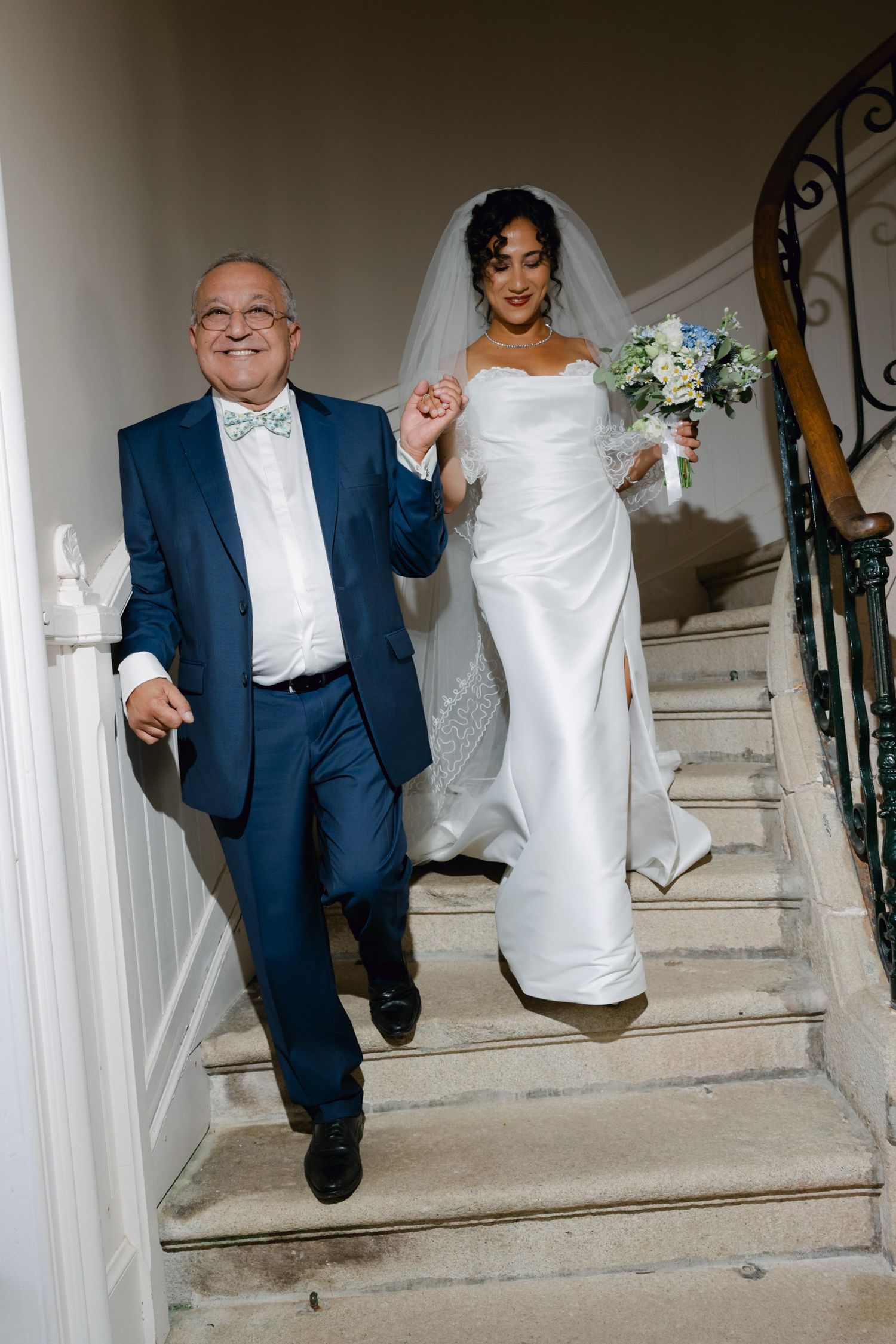 Bride in strapless white gown descends staircase holding hands with man in blue suit and bow tie.