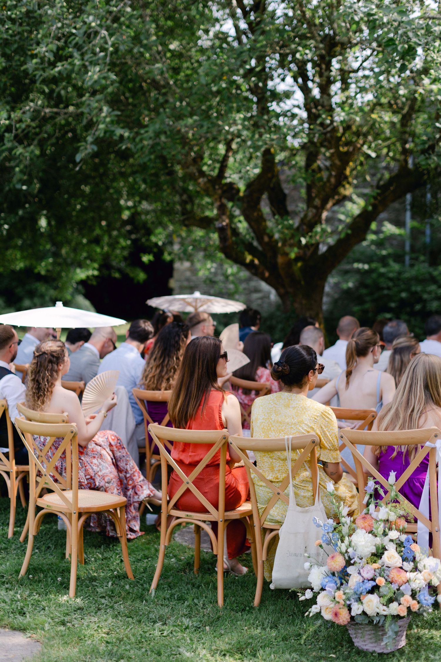 Des invités assis sur des chaises croisées en bois lors d'une cérémonie de mariage dans un jardin en plein air avec des arrangements floraux