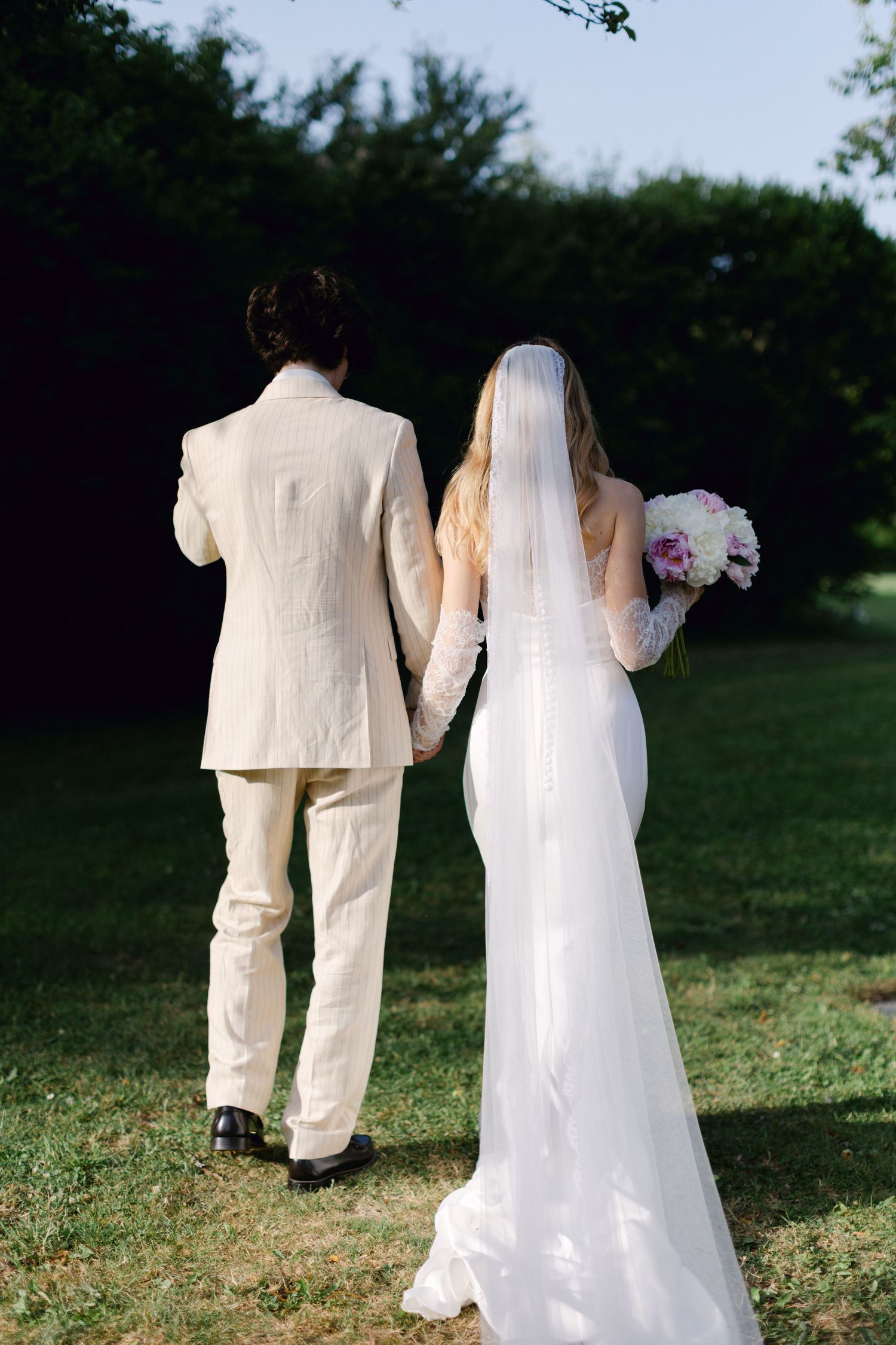 La mariée et le marié marchent main dans la main à travers un jardin verdoyant au coucher du soleil.