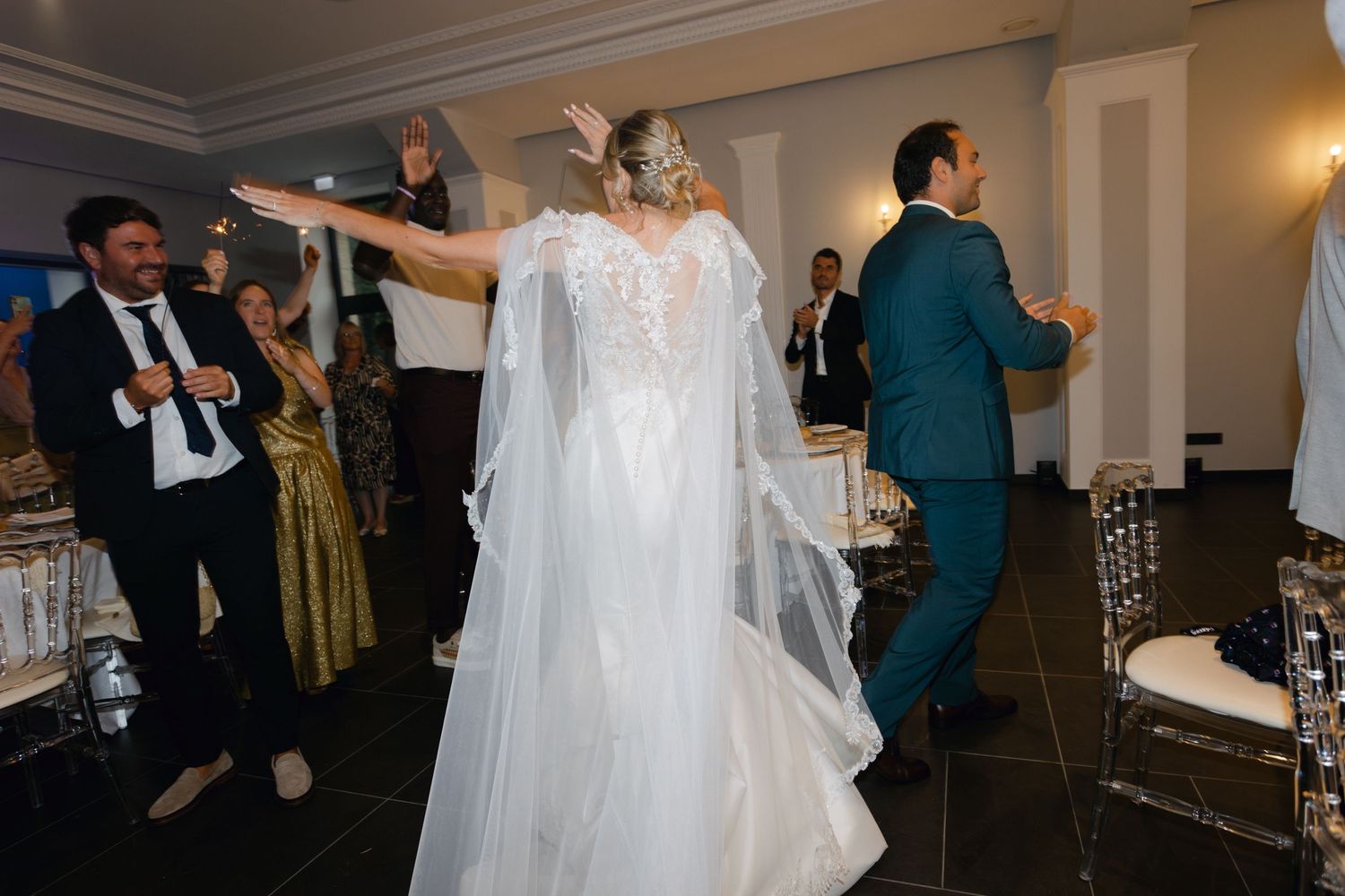 La mariée en robe cape en dentelle et le marié en costume sarcelle dansent énergiquement à la réception de mariage.