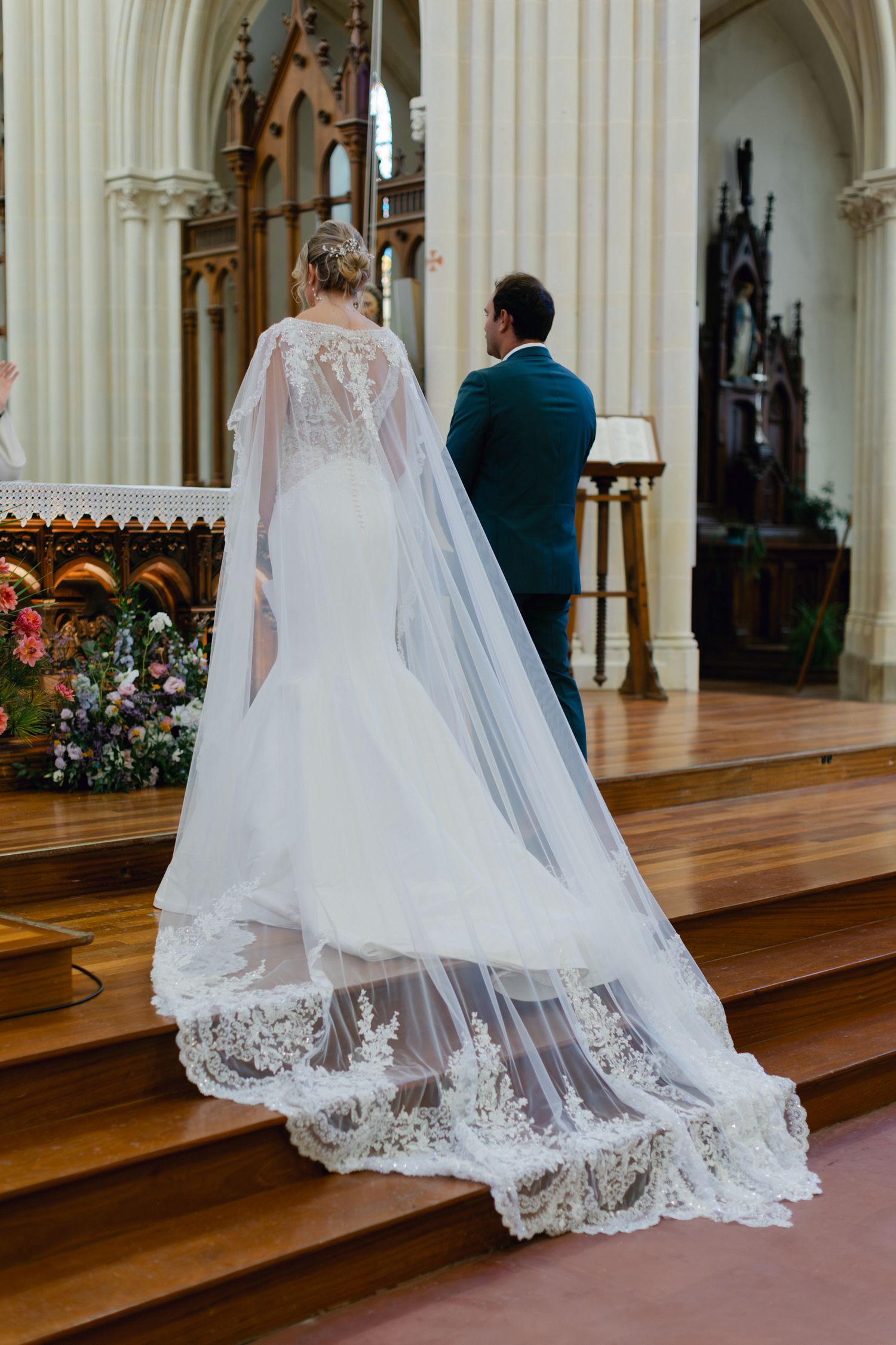 La mariée en robe de sirène en dentelle avec une cape de voile de cathédrale se tient à l'autel de l'église avec le marié pendant la cérémon