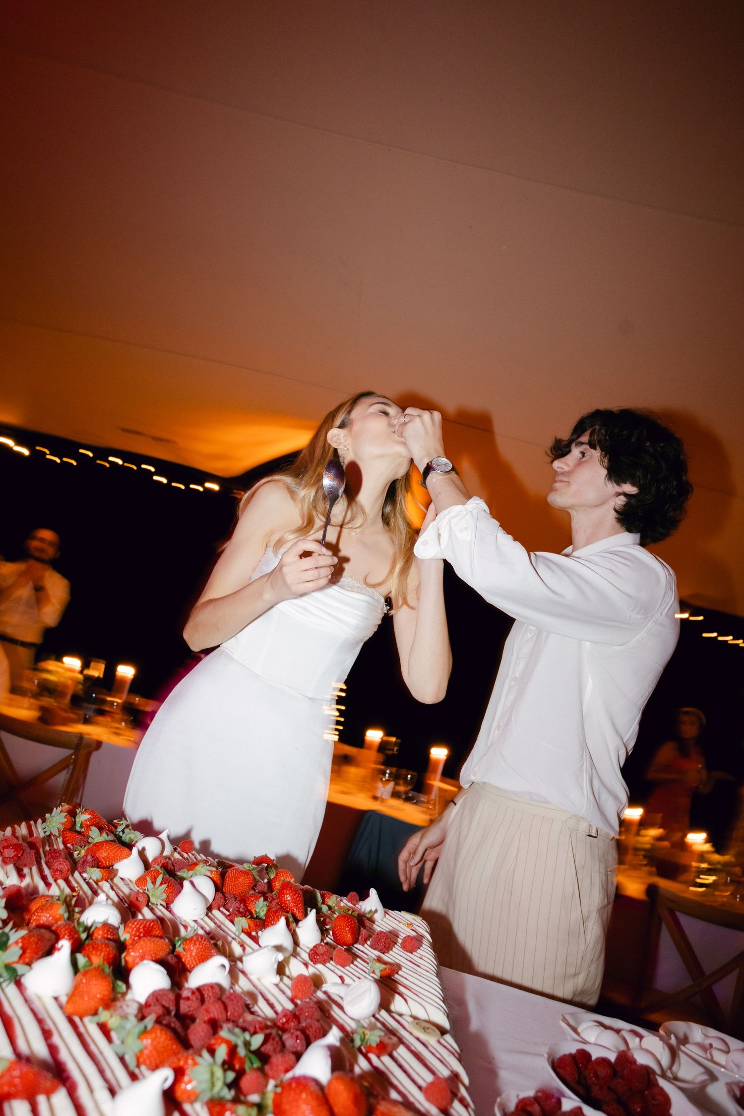 La mariée rit alors que le marié nourrit son gâteau à la réception de mariage avec une table de dessert à la fraise.