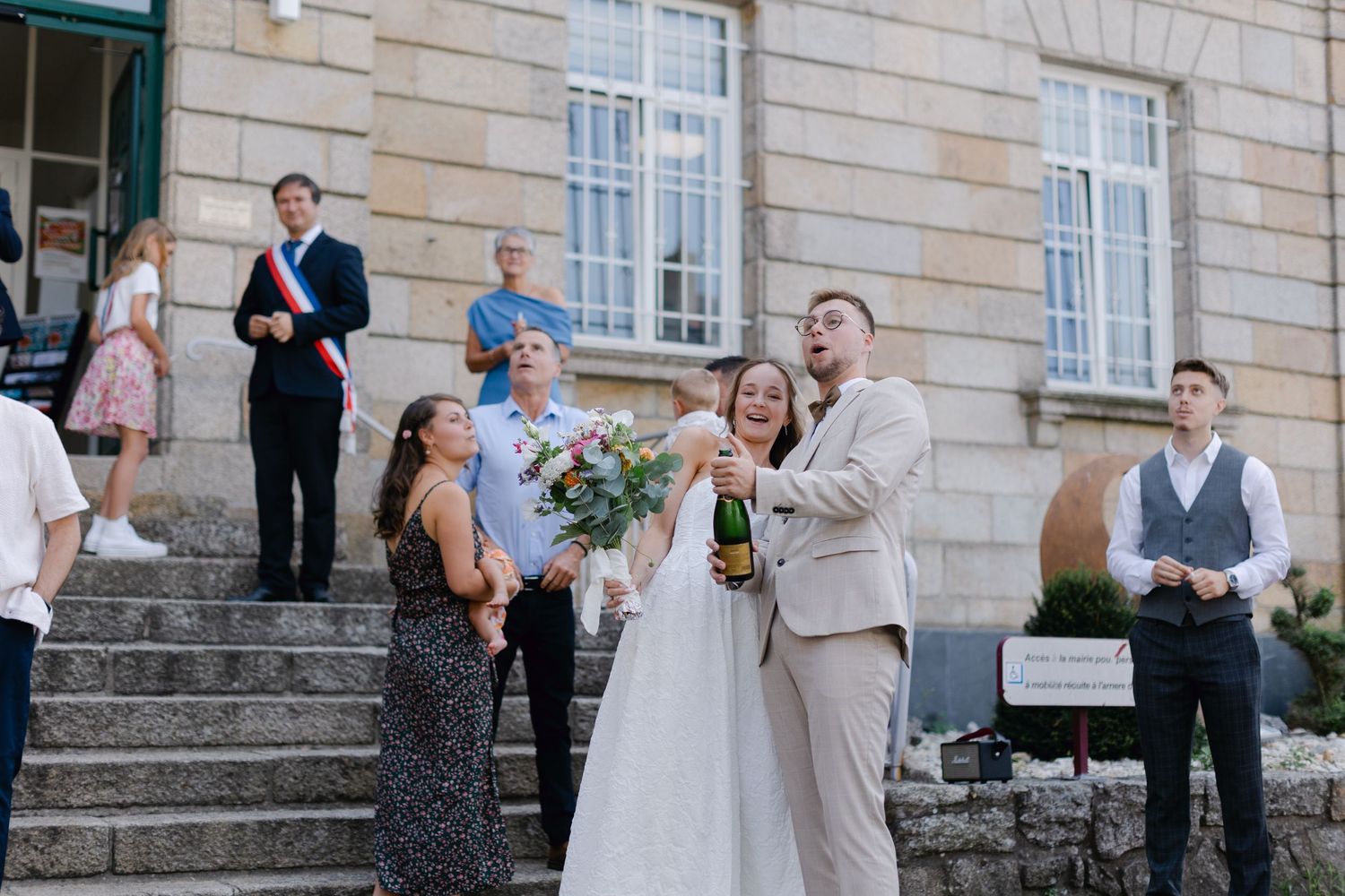 De joyeux jeunes mariés jettent une bouteille de champagne à l'extérieur du French Hotel de Ville entourés d'invités de mariage.