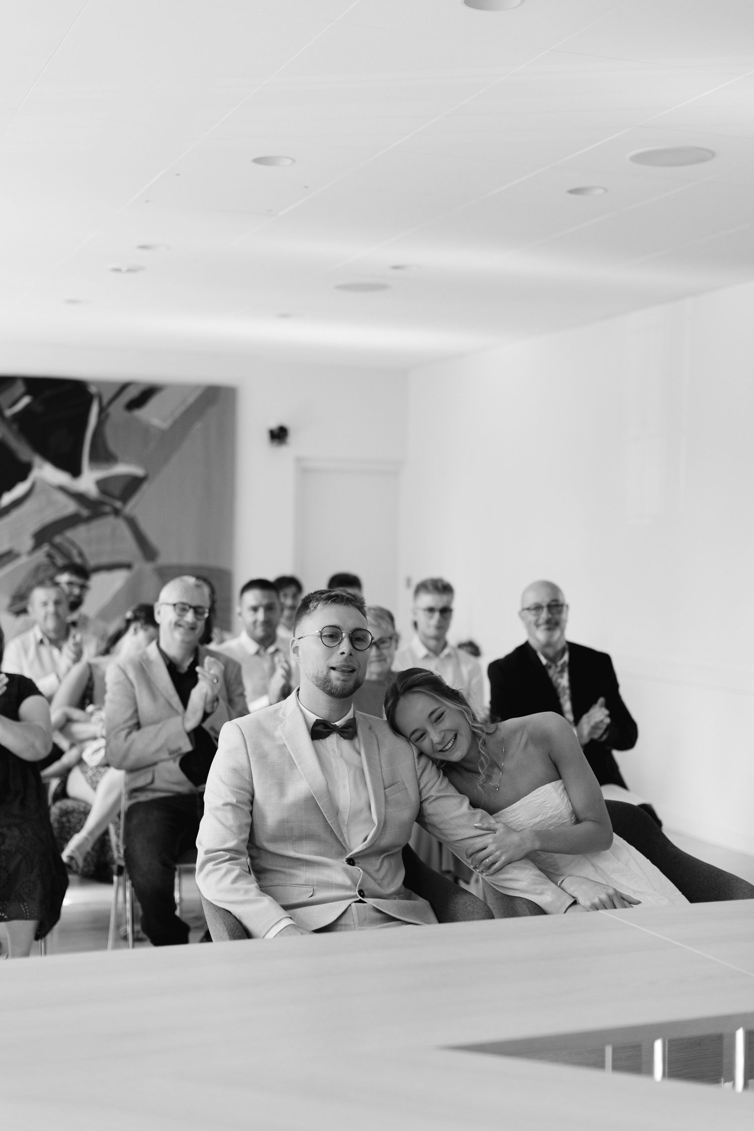 Photo en noir et blanc de la mariée appuyée sur le marié lors d'une cérémonie de mariage civil avec des invités applaudissant.