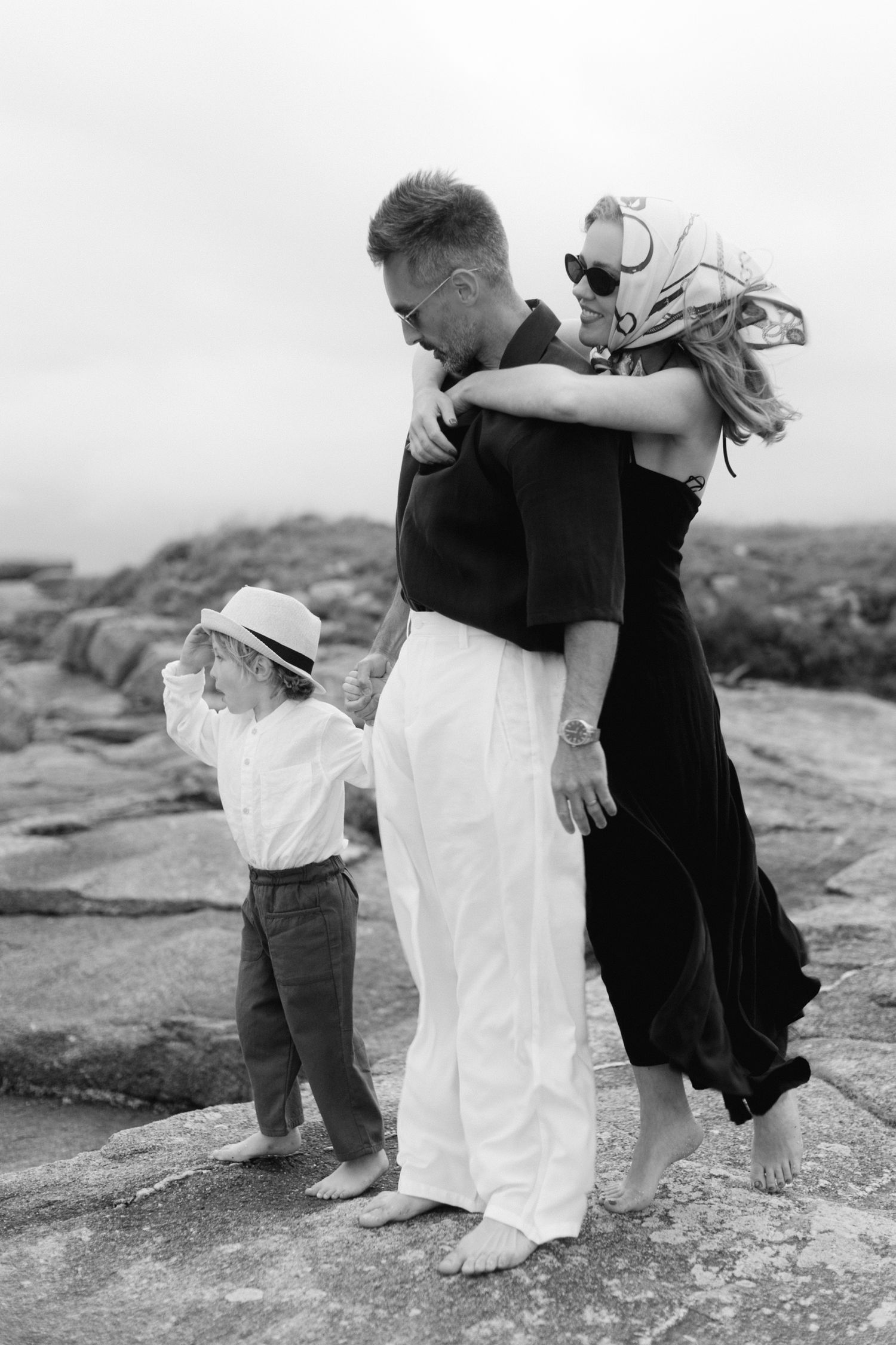 Photo en noir et blanc d'une famille sur une plage rocheuse, un couple s'embrassant tandis qu'un jeune enfant au chapeau se tient à côté d'e