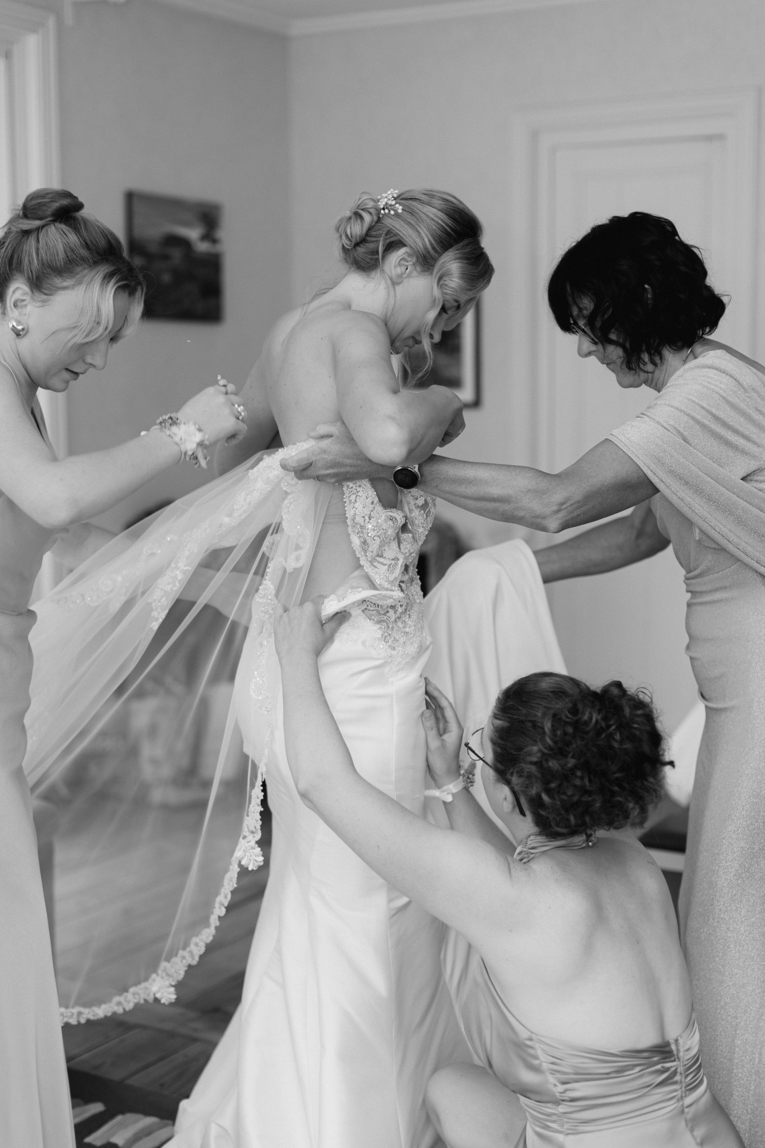 Photo en noir et blanc de demoiselles d'honneur aidant la mariée à attacher sa robe de mariée en dentelle à l'intérieur.