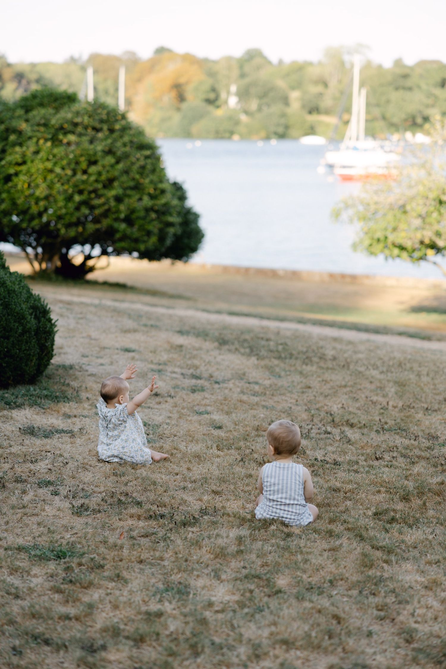 Two toddlers sitting on dry grass near a waterfront park with sailboats and lush green trees in the background.
