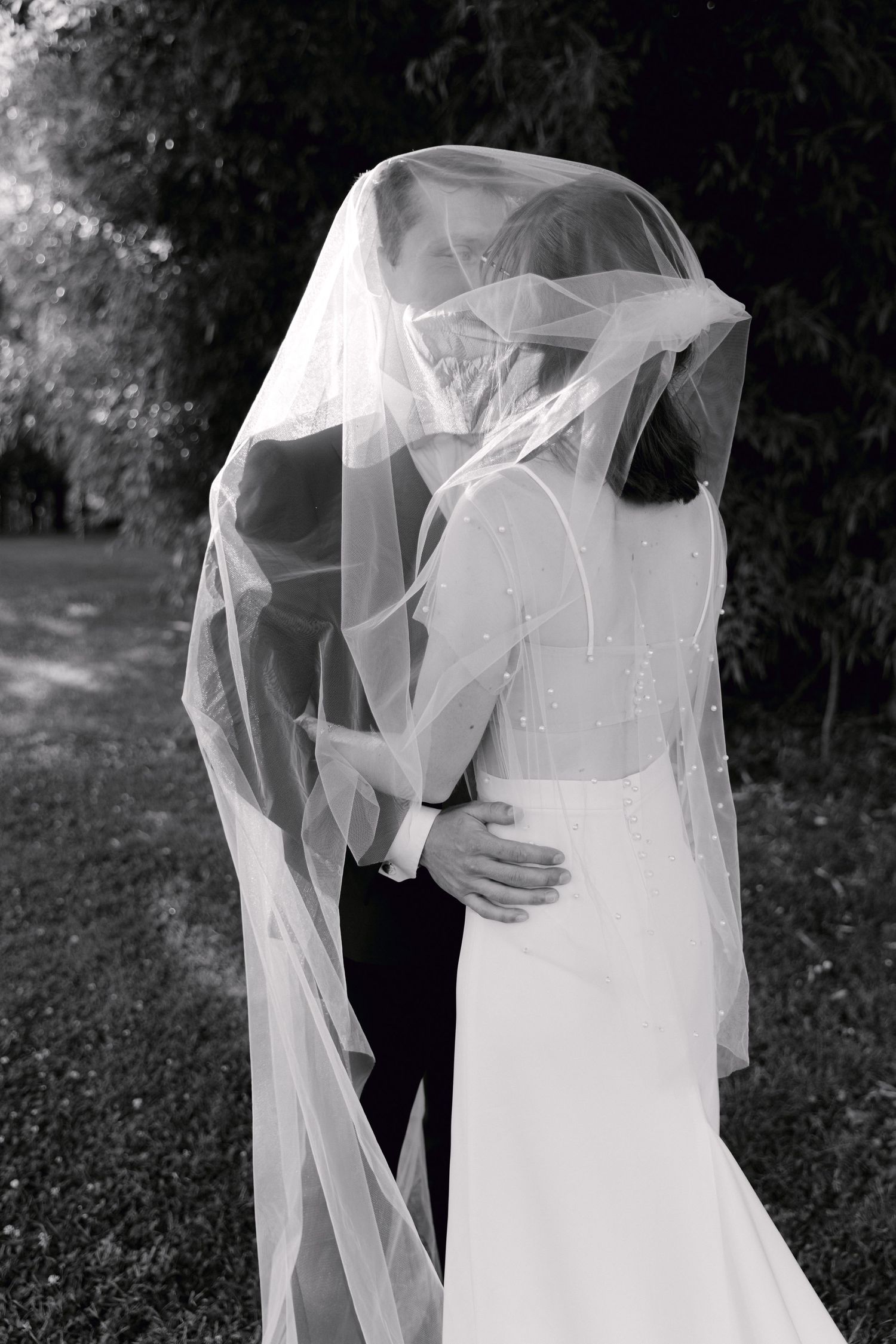 Photo en noir et blanc de la mariée et du marié s'embrassant sous un voile de mariage fluide à l'extérieur.