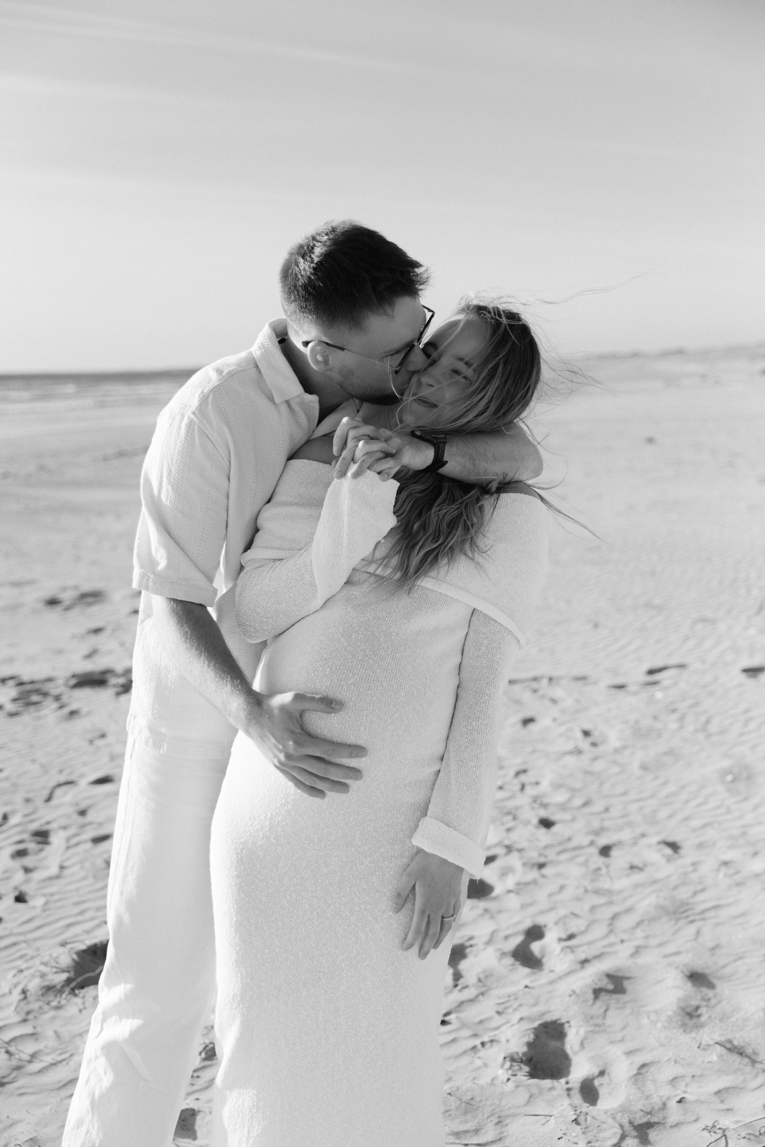 Photo en noir et blanc d'un homme embrassant une femme enceinte sur une plage de sable, tous deux en tenues blanches.