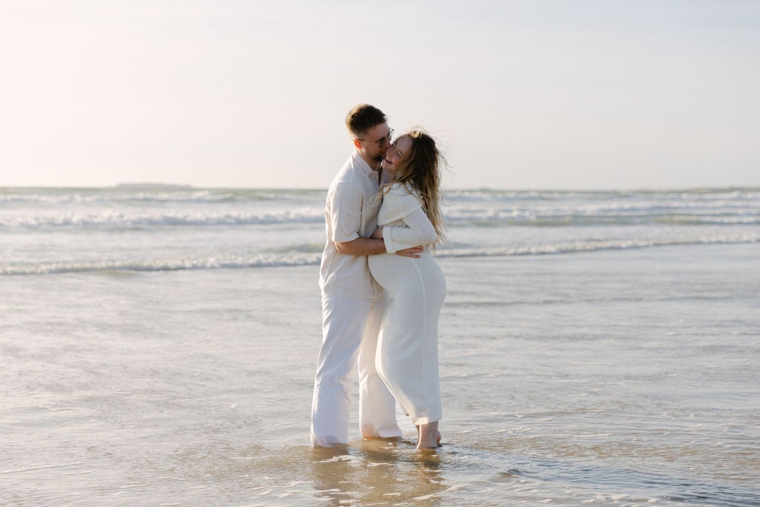 Couple heureux en tenues blanches s'embrassant et s'embrassant tout en se tenant dans l'eau peu profonde de l'océan sur une plage.