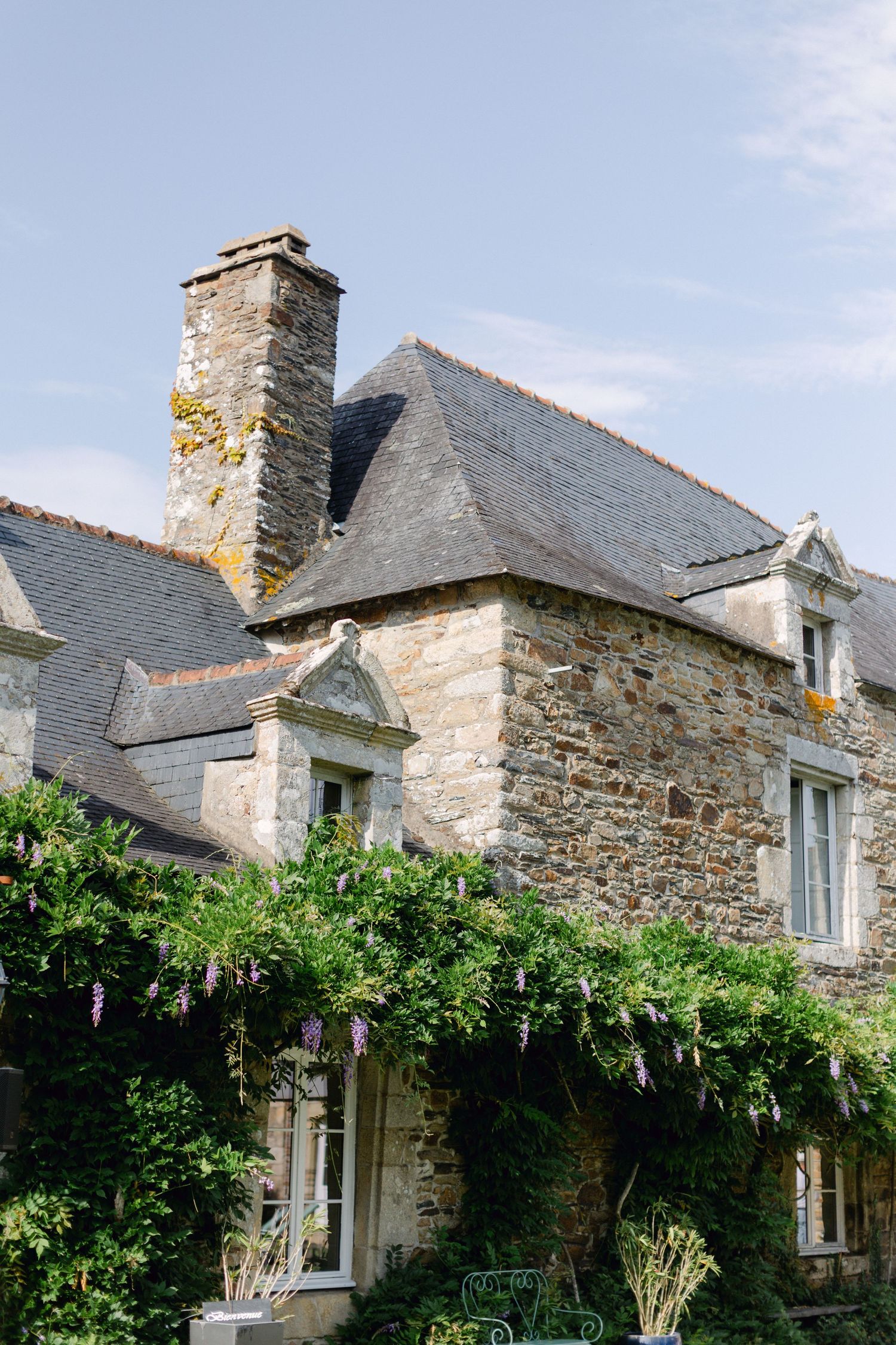 Manoir historique en pierre avec toit en ardoise et glycine grimpant sur la façade sous un ciel bleu.
