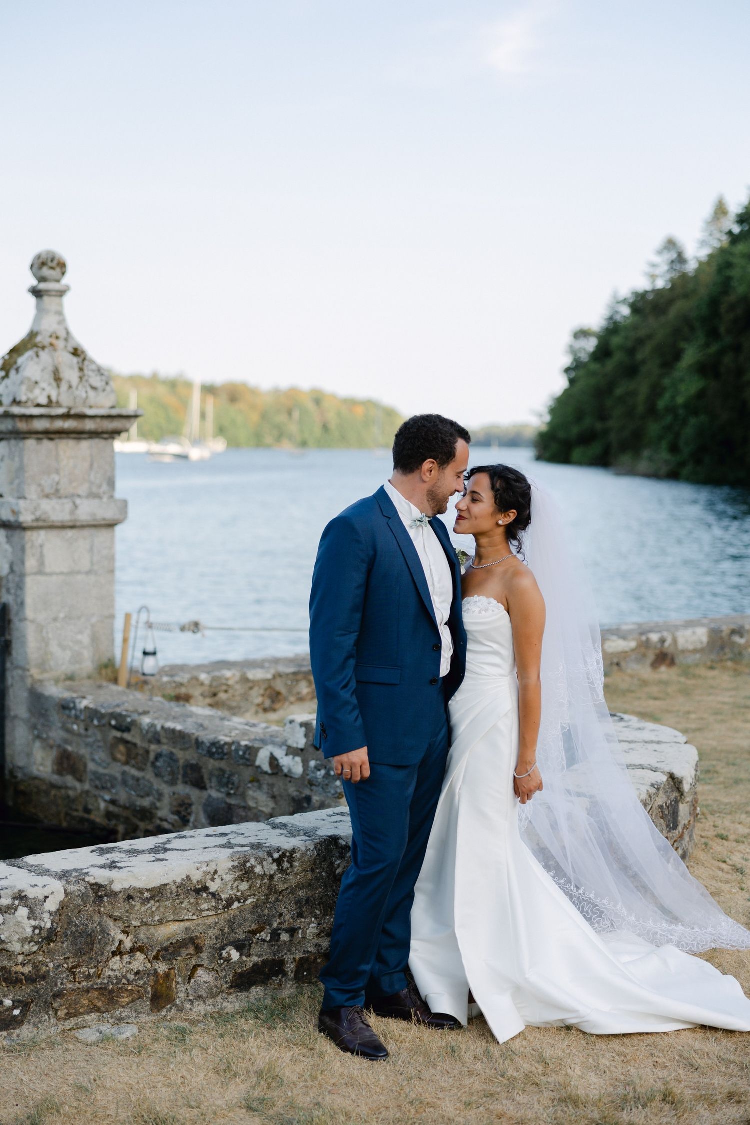 Bride and groom share a romantic moment by a waterside stone wall, with lush trees and calm water in the background.