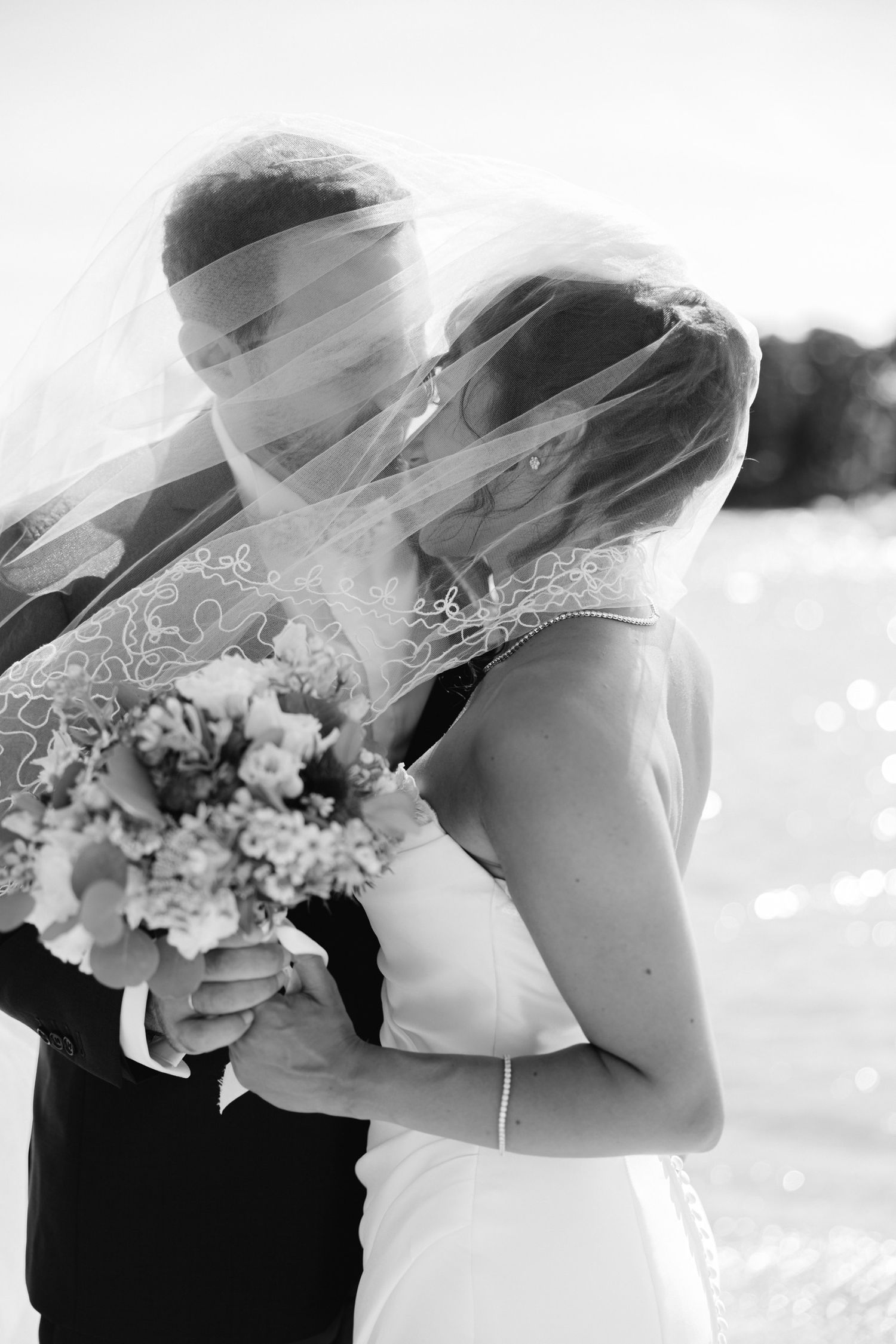 Black and white photo of bride and groom kissing under wedding veil holding floral bouquet outdoors.