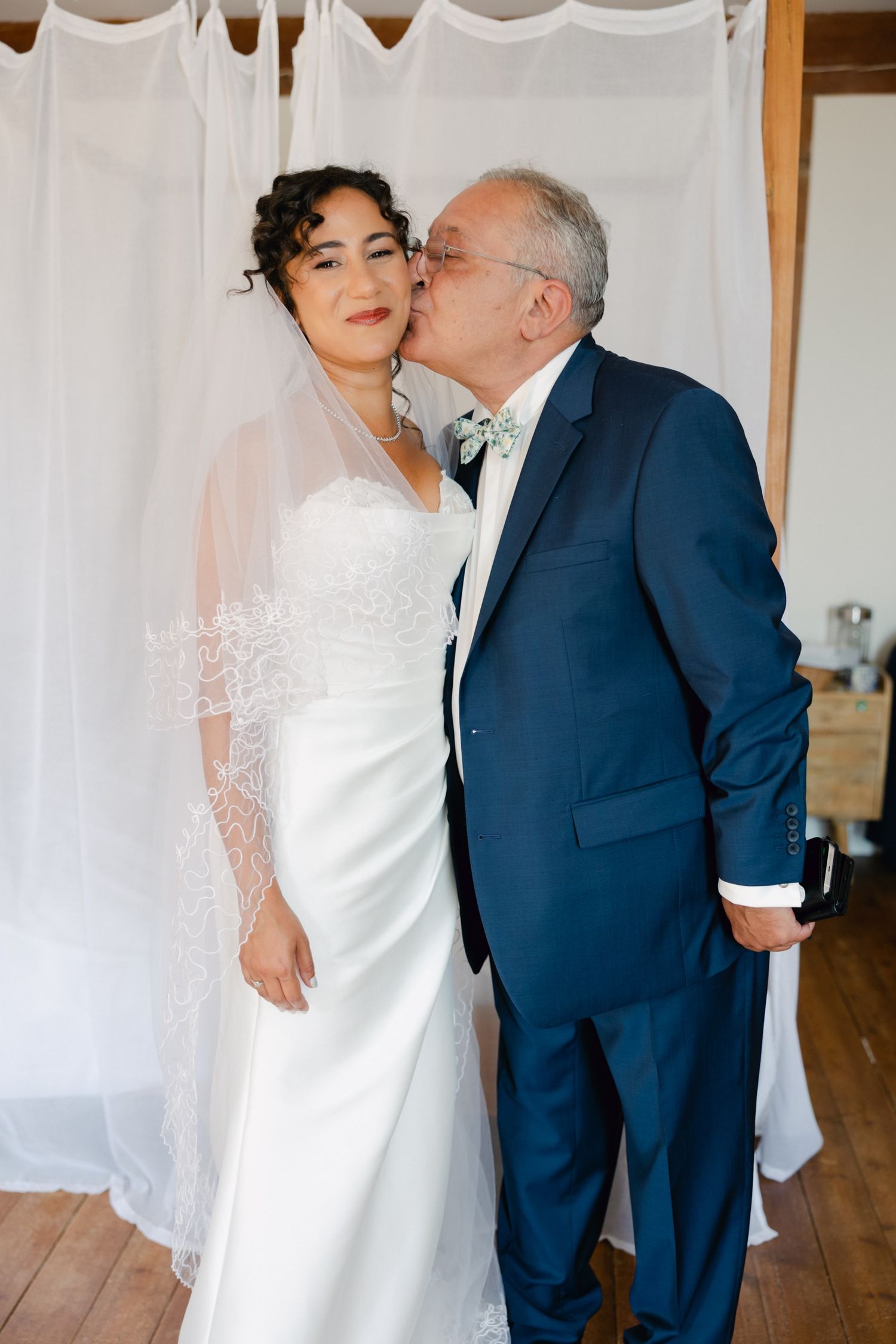 Bride in white strapless gown and veil receives a kiss on the cheek from an older man in a navy suit.