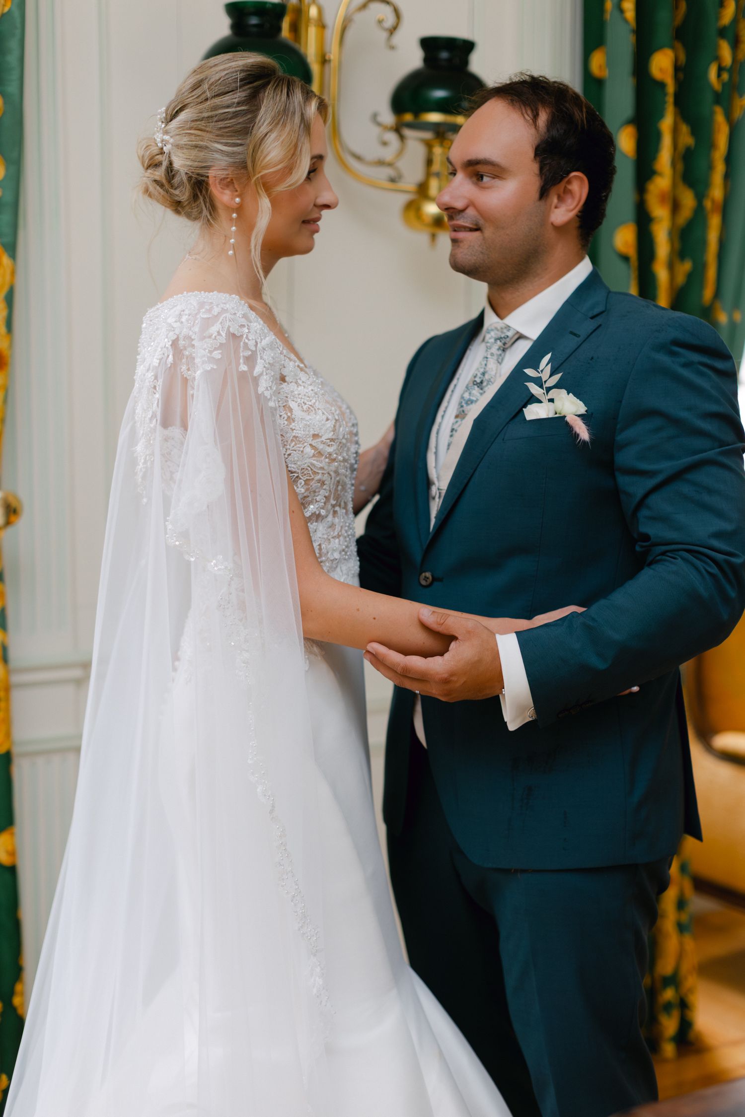 La mariée et le marié se tenant la main partageant un moment romantique dans une chambre ornée avec des rideaux verts.