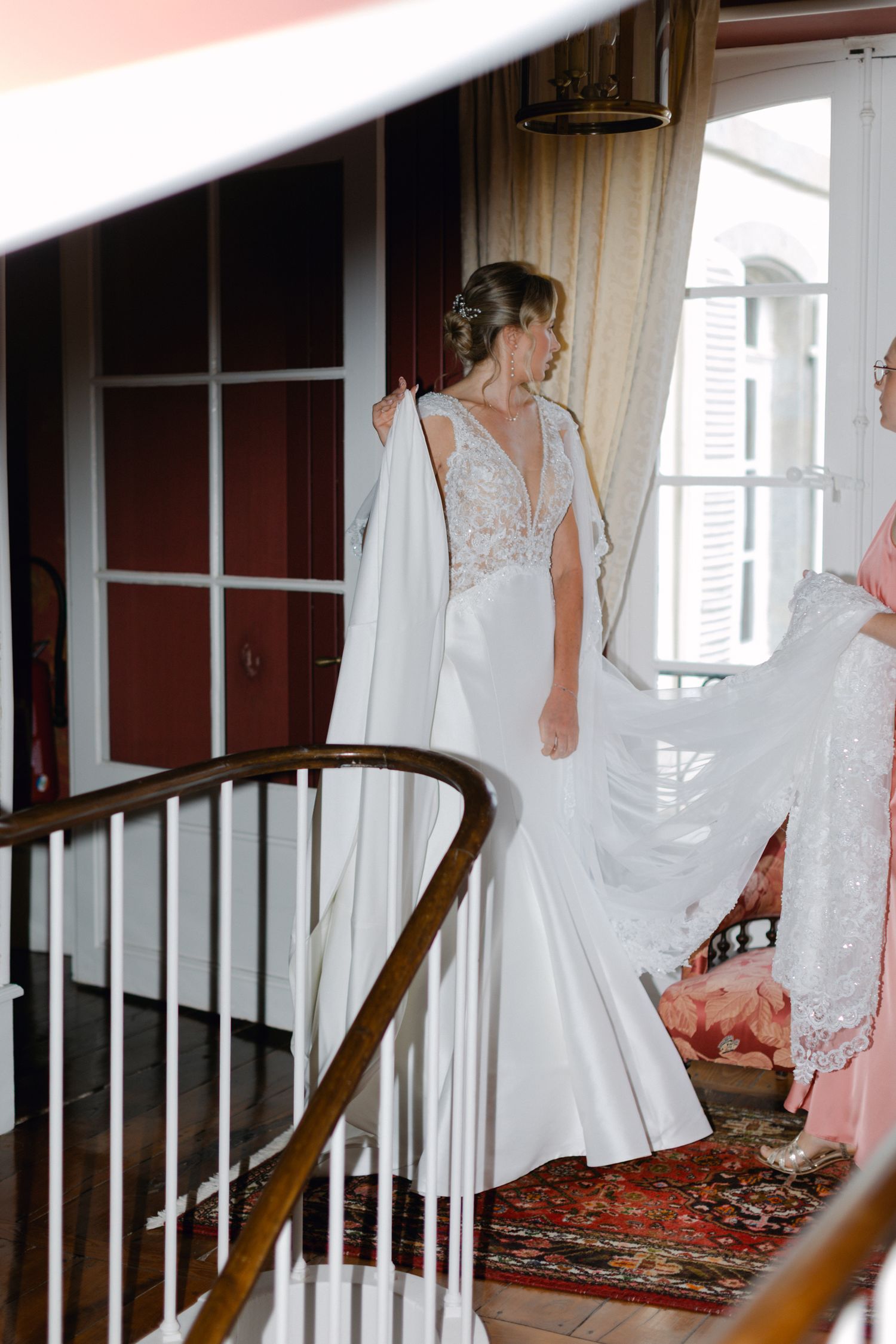 La mariée en robe de sirène en dentelle blanche avec voile de cape descend les escaliers assistée par une demoiselle d'honneur dans un lieu