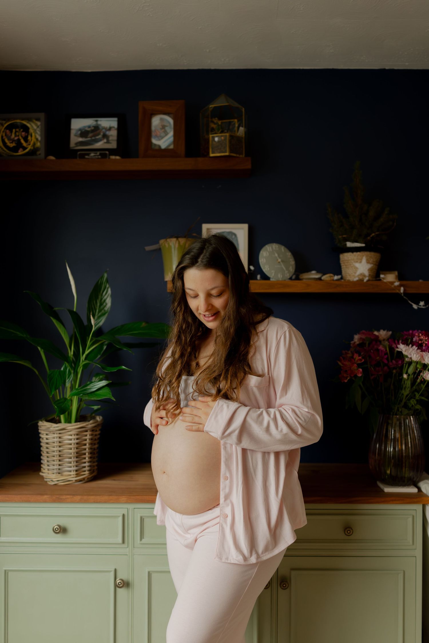 Smiling pregnant woman in pink outfit cradling her bare baby bump in a stylish navy blue kitchen.