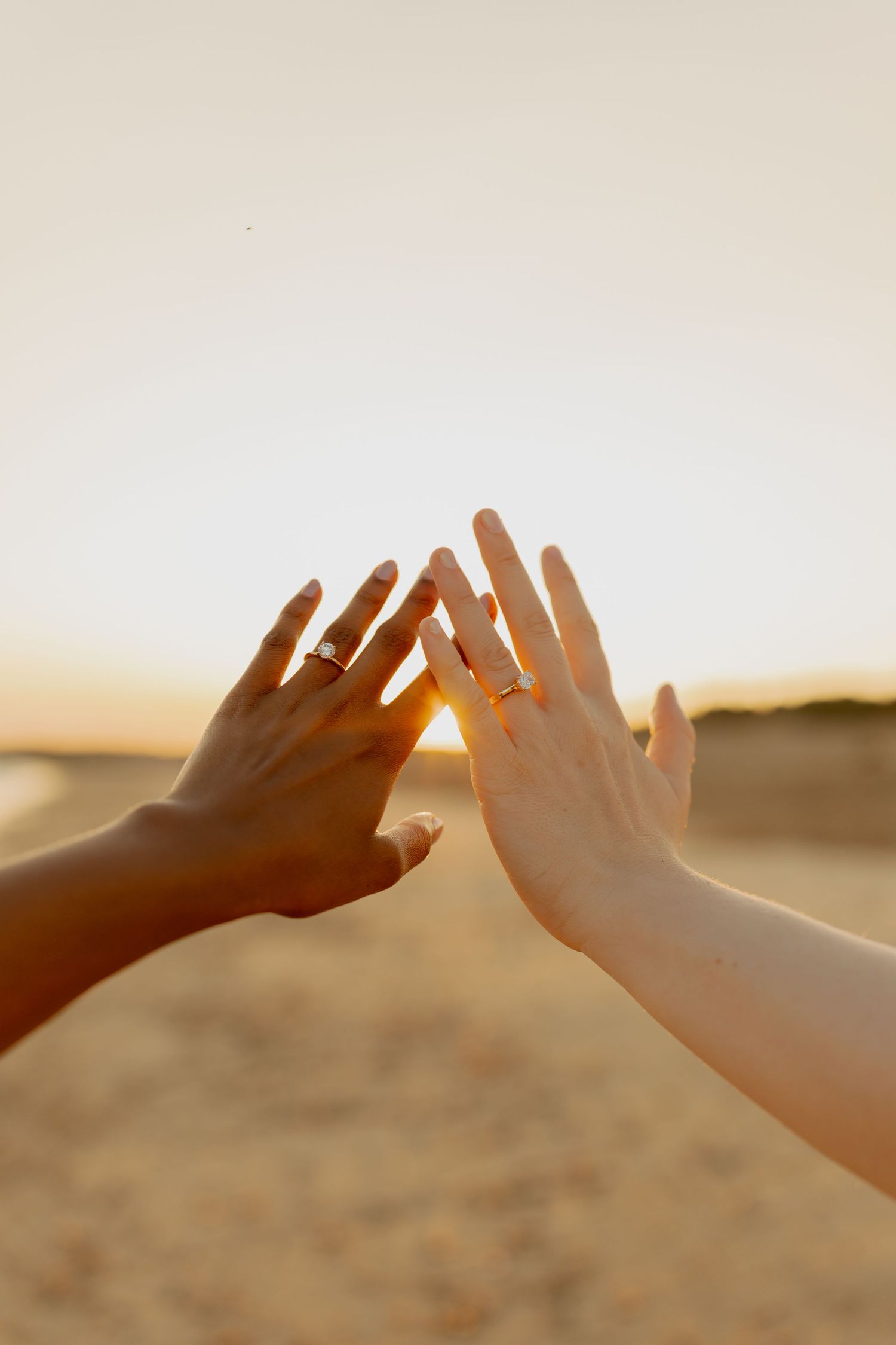 Two hands with matching rings touching at sunset on a beach, symbolizing love and commitment.