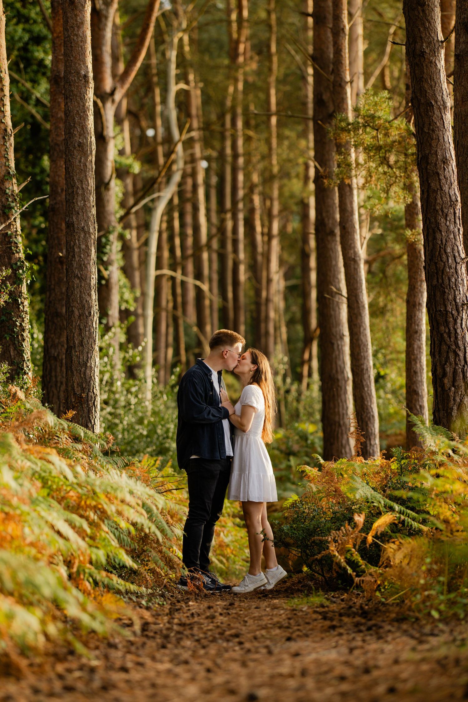 A couple shares a romantic moment on a forest path surrounded by tall pine trees.