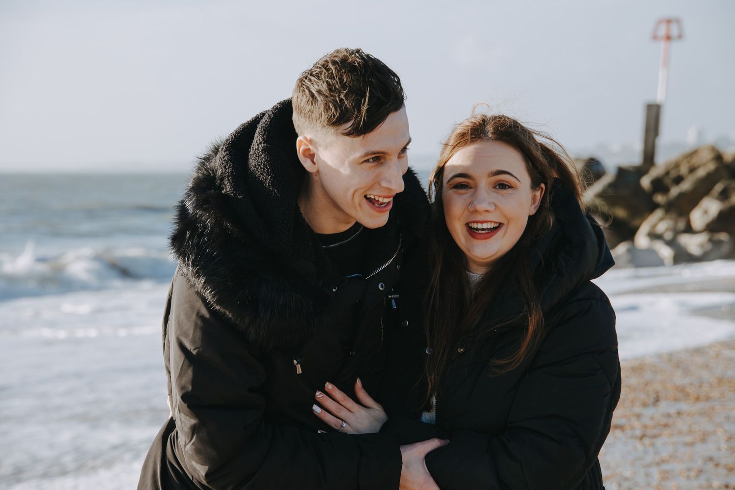A joyful couple laughing together on a rocky beach with waves crashing behind them.