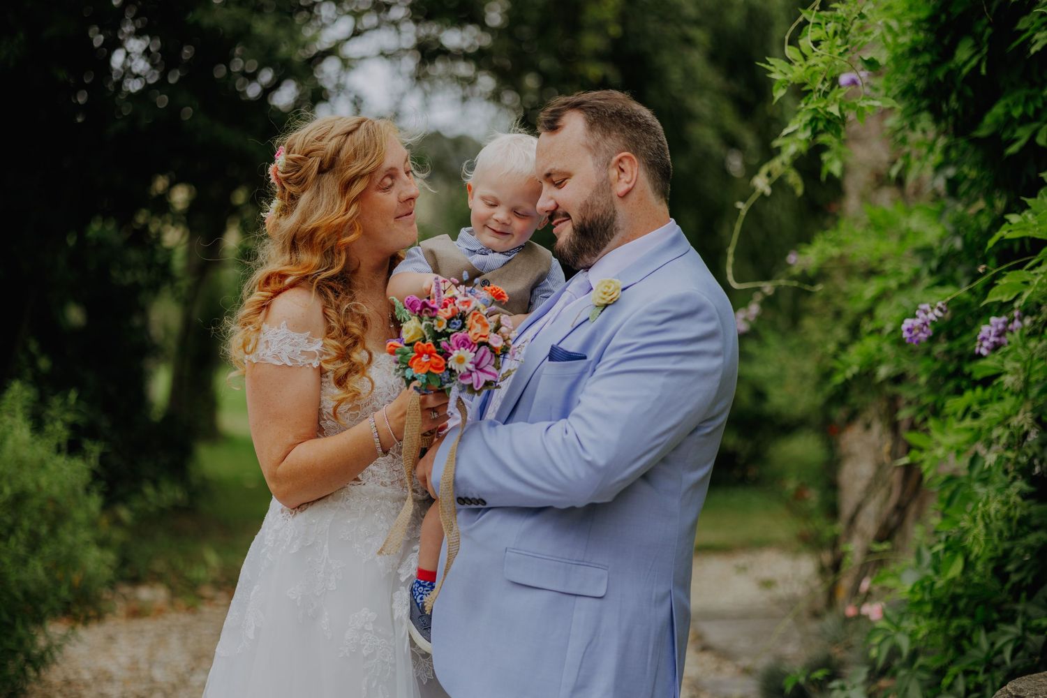Wedding couple in blue suit and white dress hold their child outdoors.