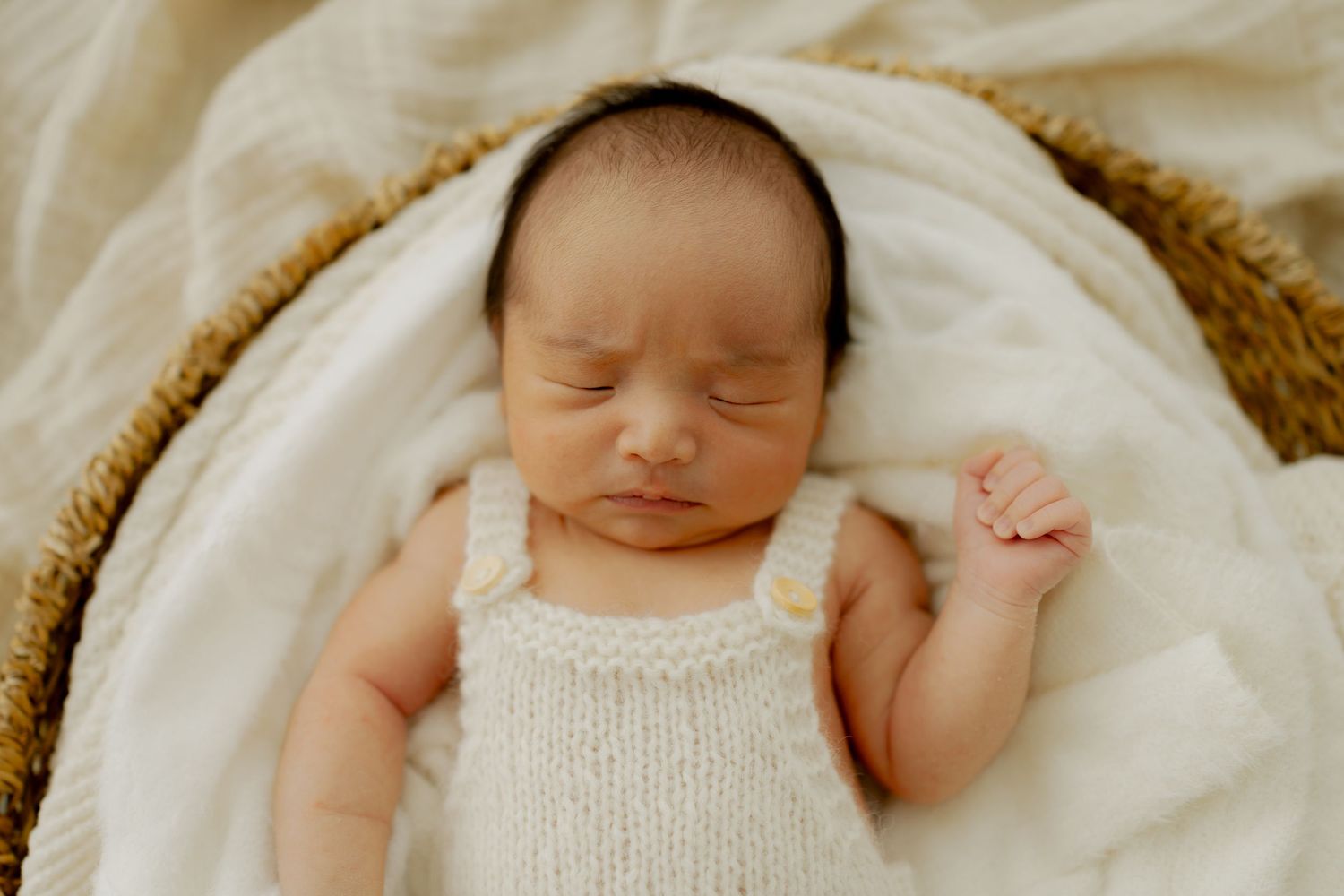 Newborn baby sleeping peacefully in a wicker basket wearing a white knit romper on a soft cream blanket.