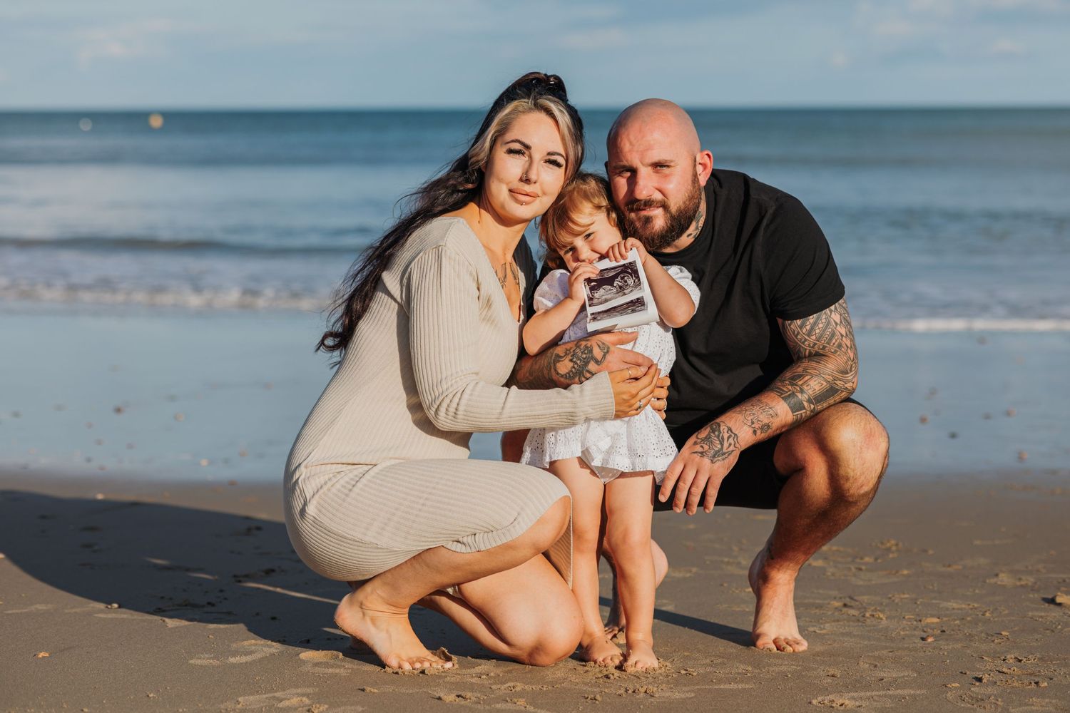 Family of three poses together on sandy beach during sunset photoshoot.