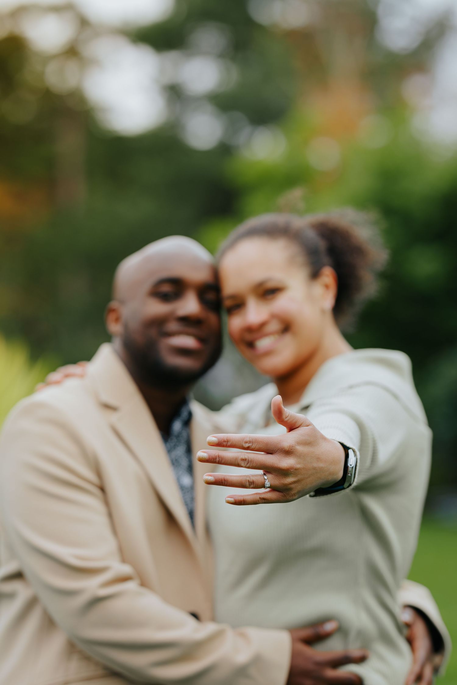 A couple shares a joyful moment while showing off an engagement ring in an outdoor garden setting.