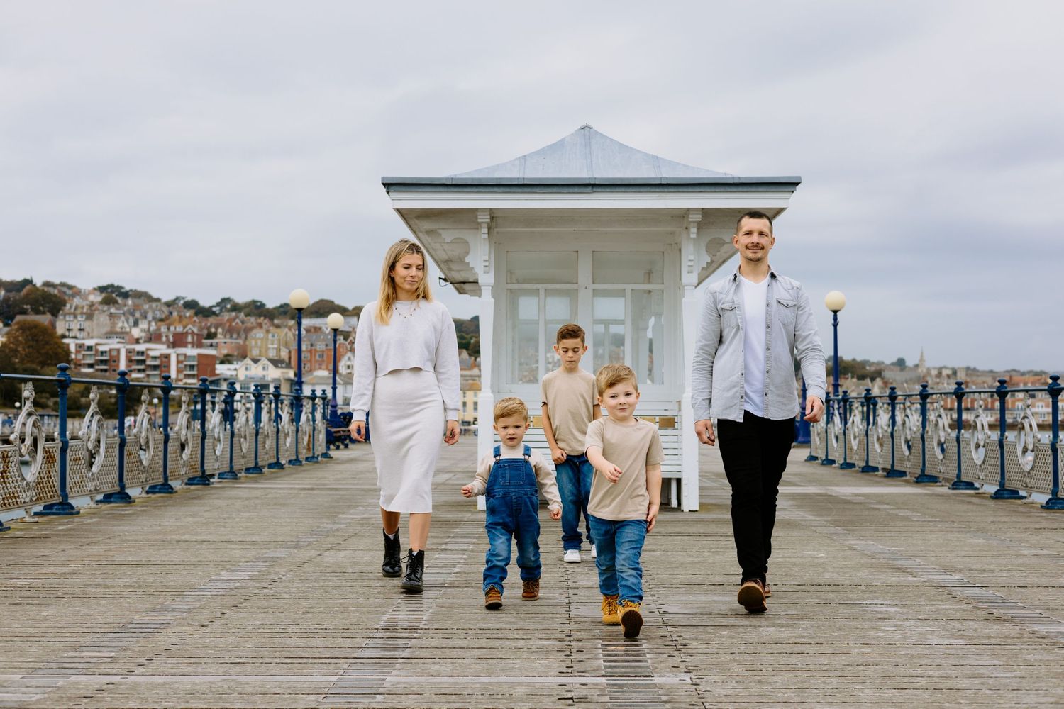 A family of five walks together on a seaside pier with a white gazebo in the background.