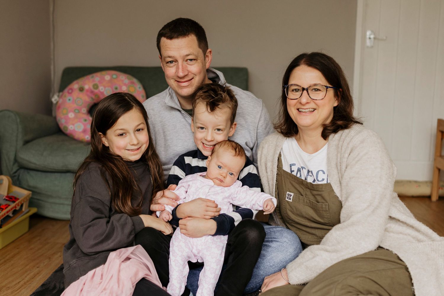 Family group sitting together on floor with infant in casual home setting with pink donut pillow visible.