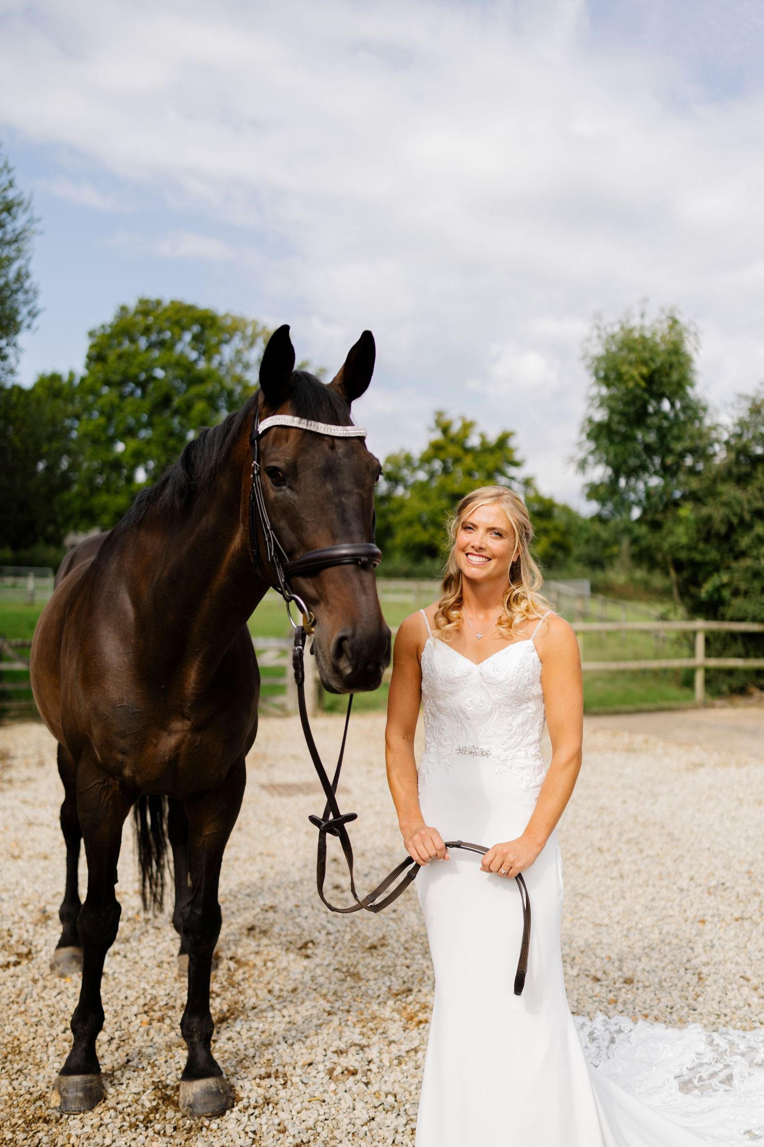 A bride in a white gown stands next to a majestic dark brown horse on a gravel path.