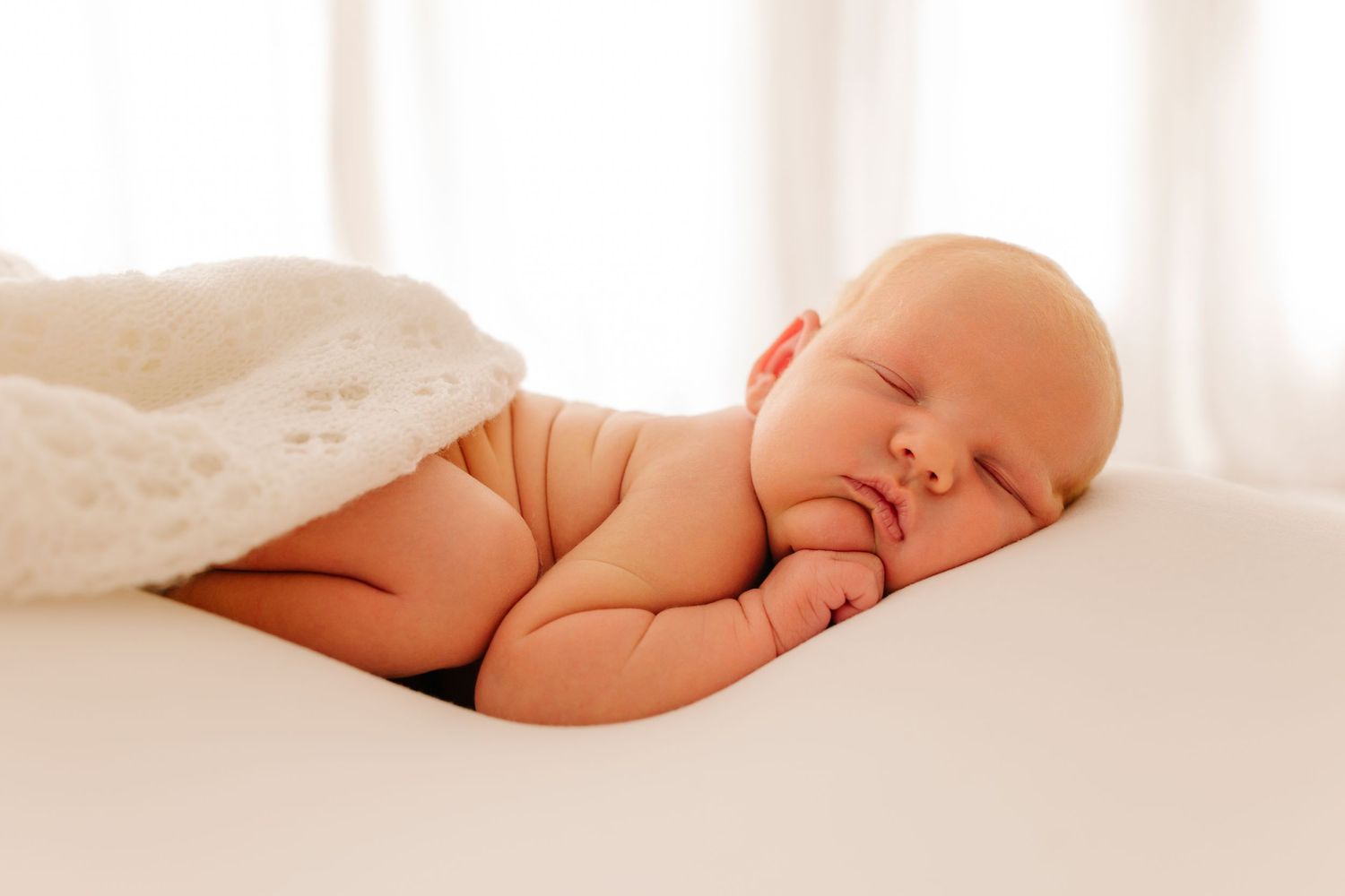 Sleeping newborn baby partially covered with white blanket against bright background.