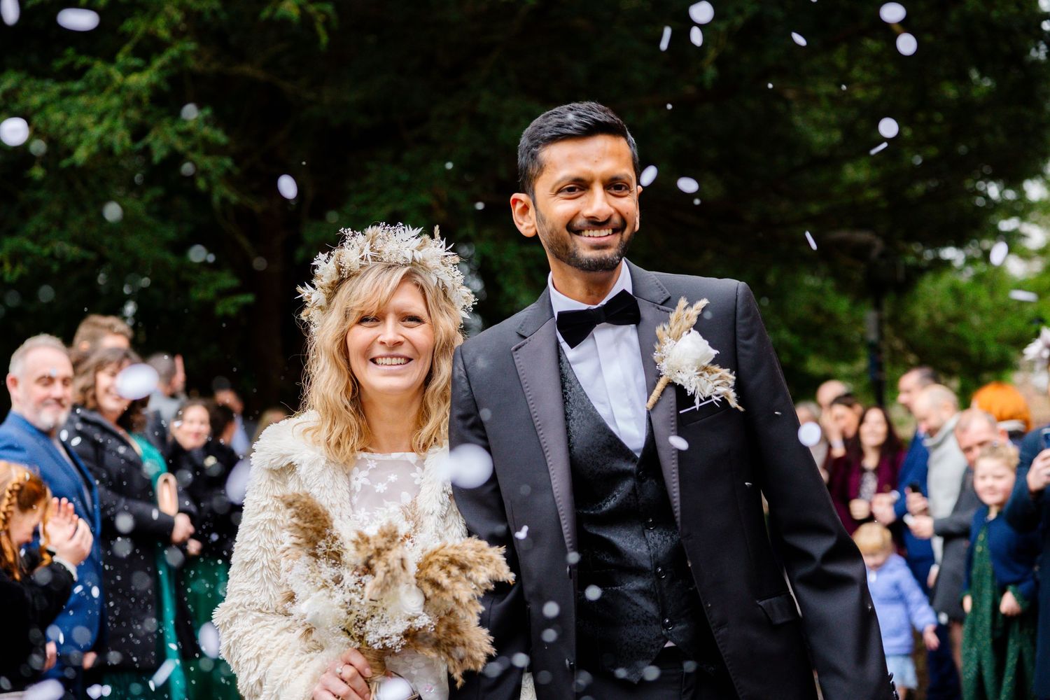 A wedding celebration outdoors with guests throwing confetti over a happy couple walking down a garden path.