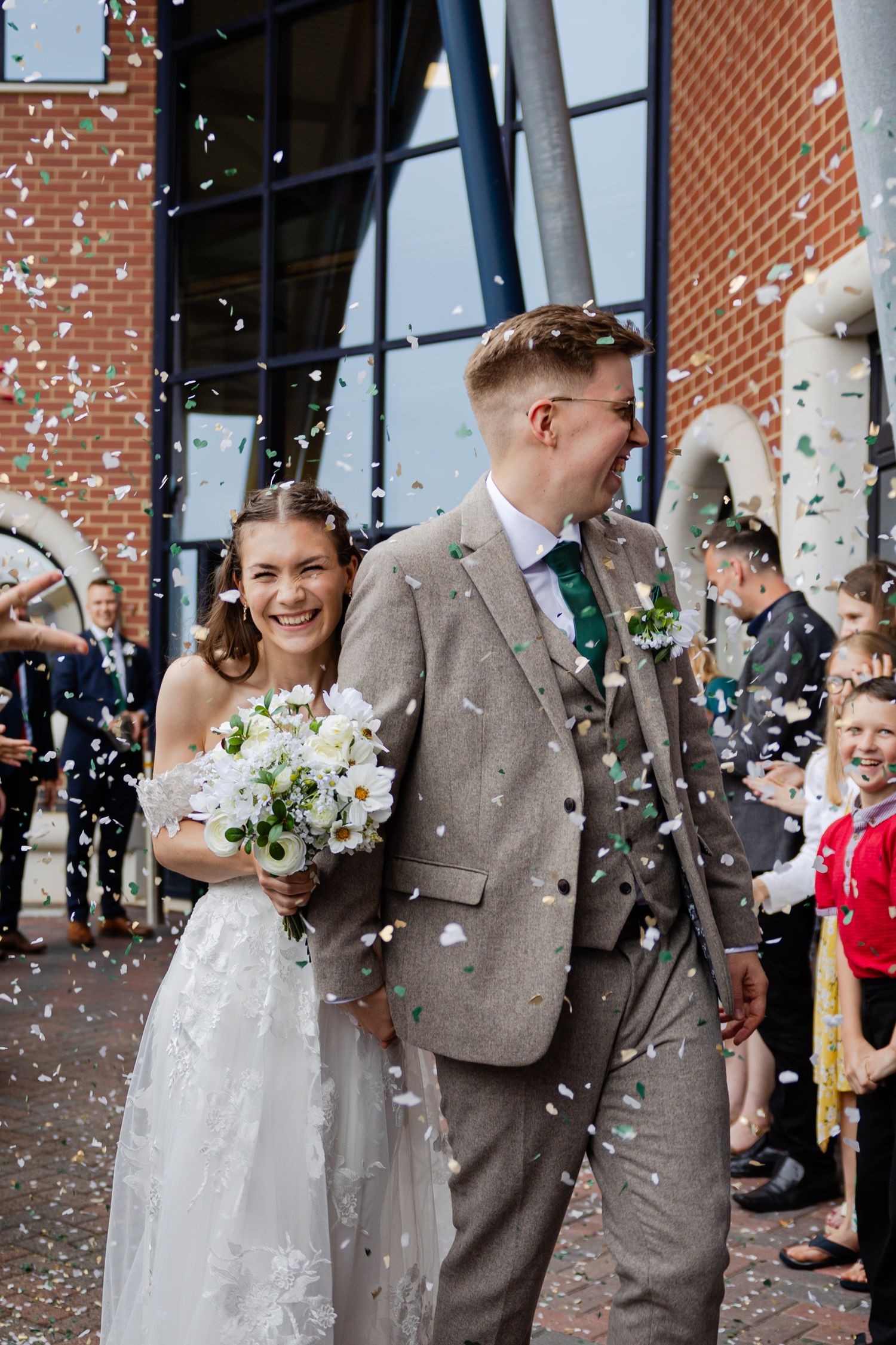 Wedding couple exits venue amid shower of confetti with brick building backdrop.