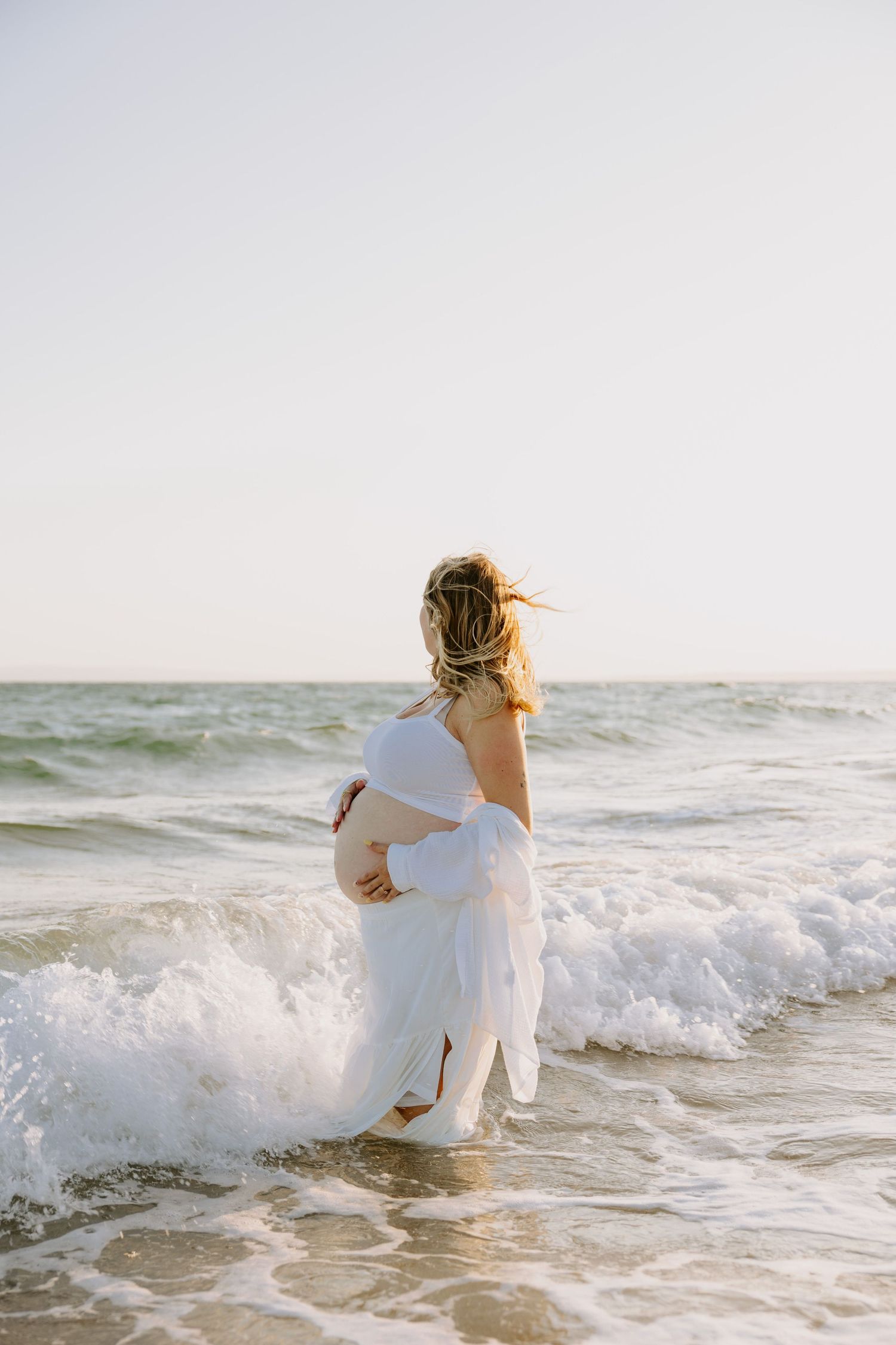 Expectant mother in white dress standing in ocean waves at sunset.
