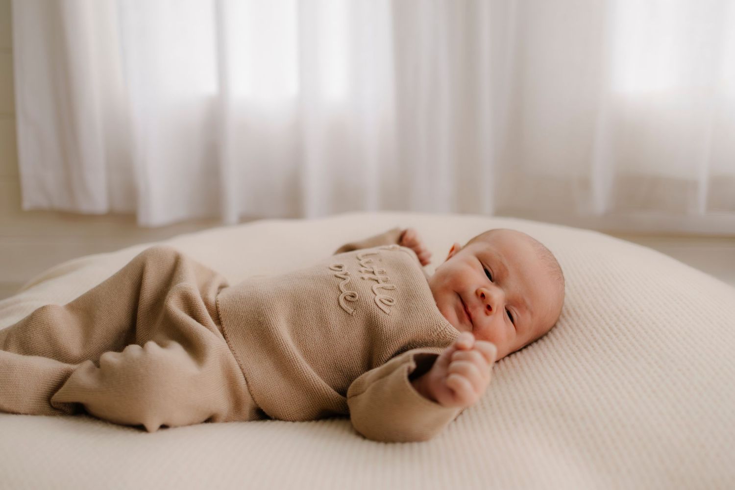 Newborn baby sleeping peacefully on white textured bedding in natural window light.