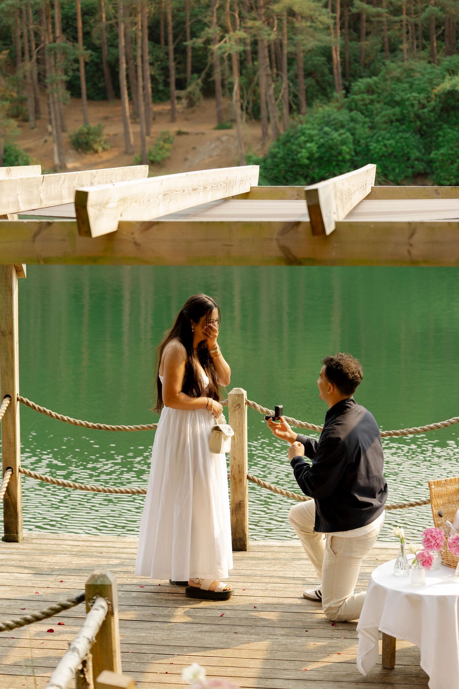 A romantic moment unfolds on a lakeside wooden dock surrounded by lush forest and green tranquil waters.