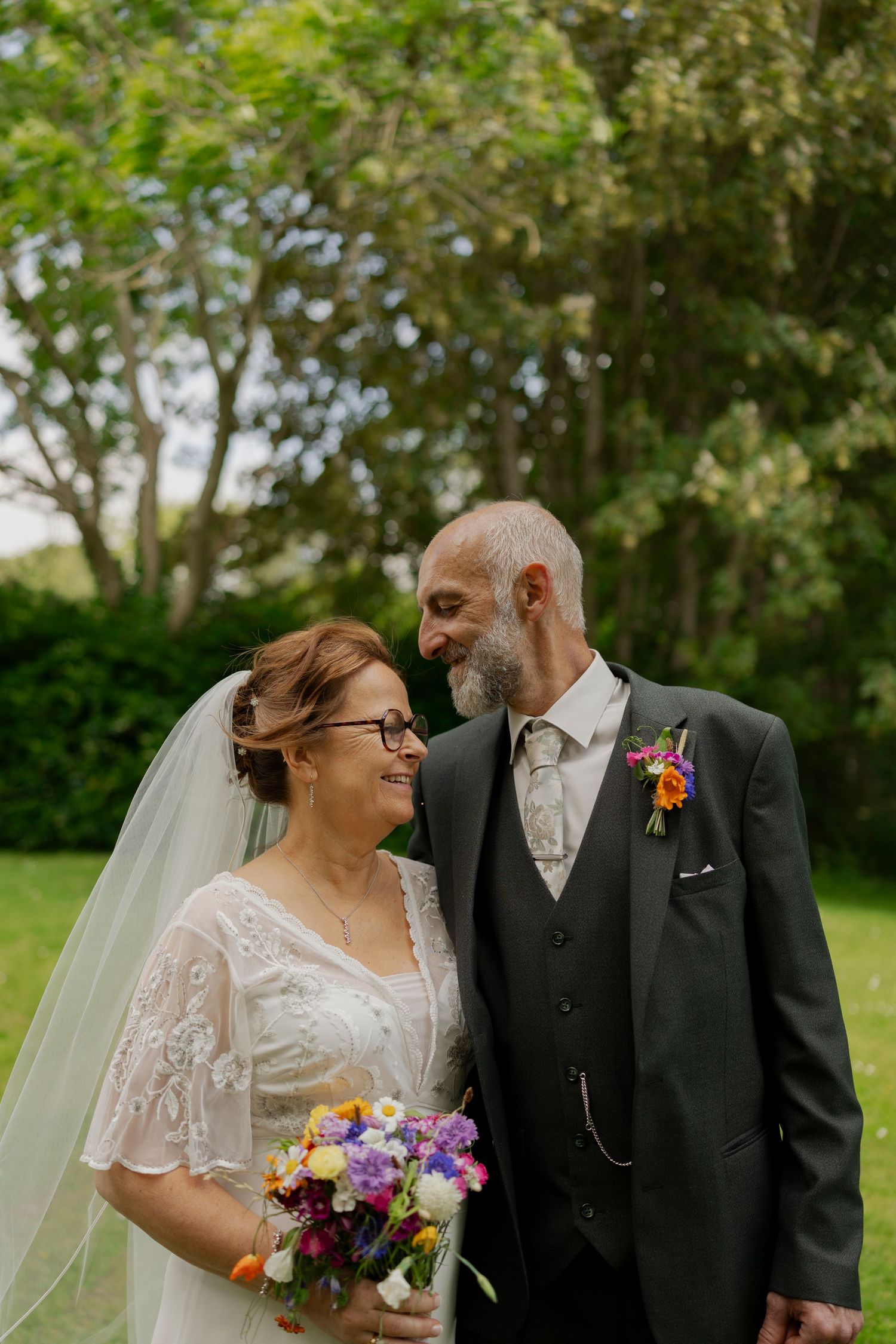 A couple in wedding attire shares happy glances and laughter while standing together in a lush outdoor garden setting.