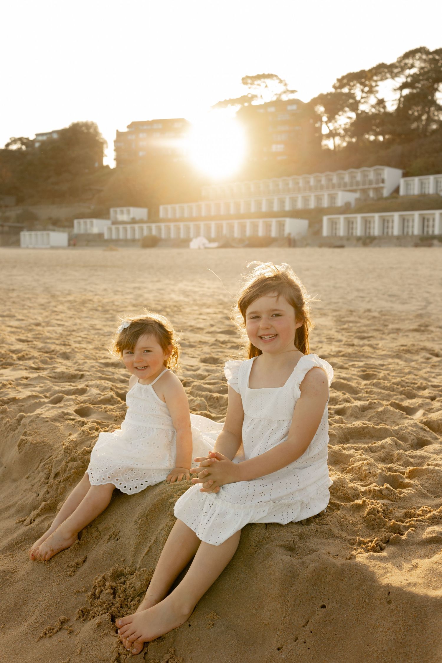 Two young girls in white dresses sitting on sandy beach at golden sunset with white buildings in background.